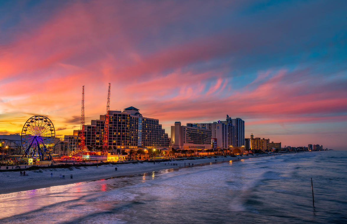 Sunset over a waterfront city skyline with lit buildings, ferris wheel, and waves under pink-orange clouds