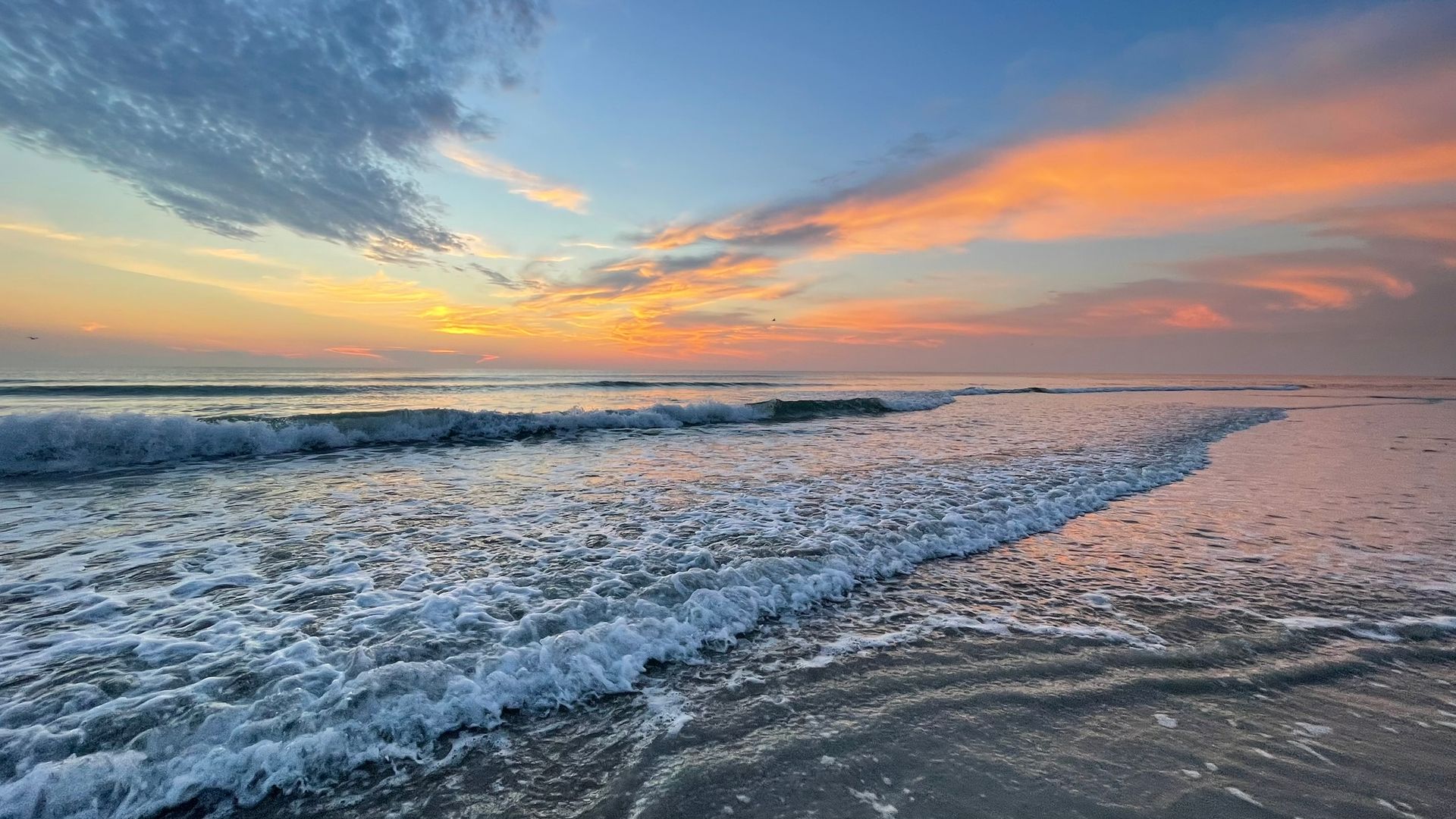 Waves rolling onto a sandy beach at sunset, with pink and orange clouds over the horizon.