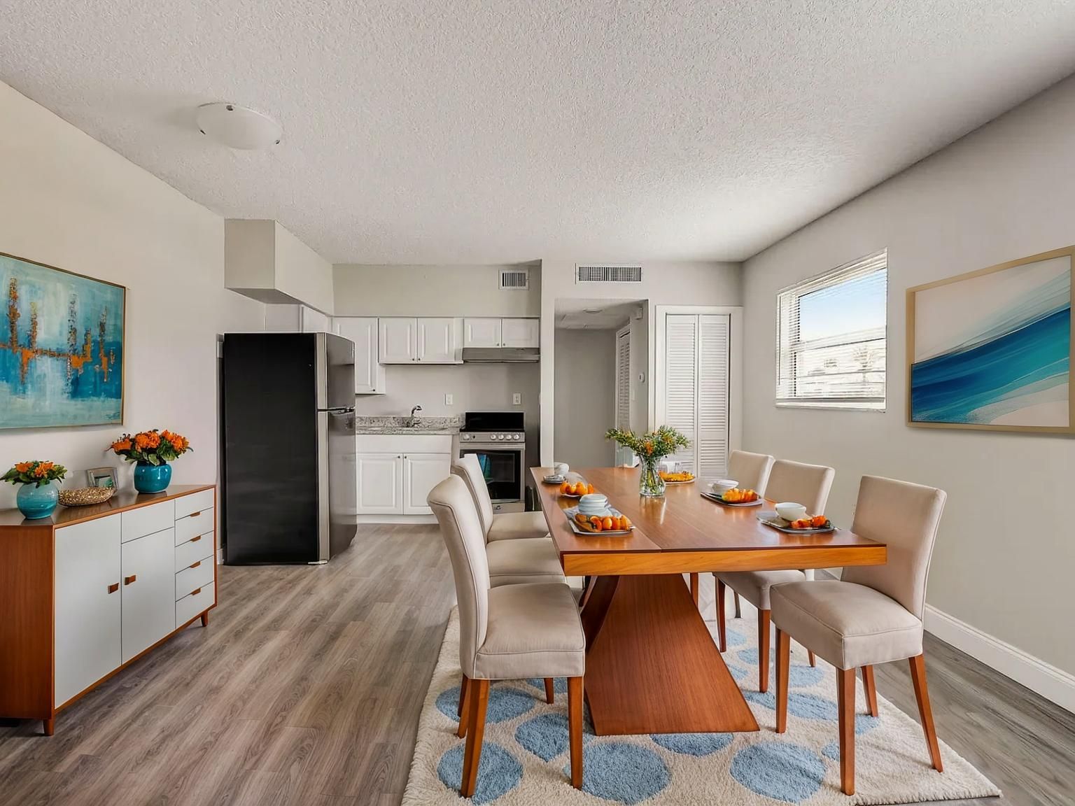 Bright dining room with wooden table set, white chairs, kitchen in back, and ocean artwork on the walls