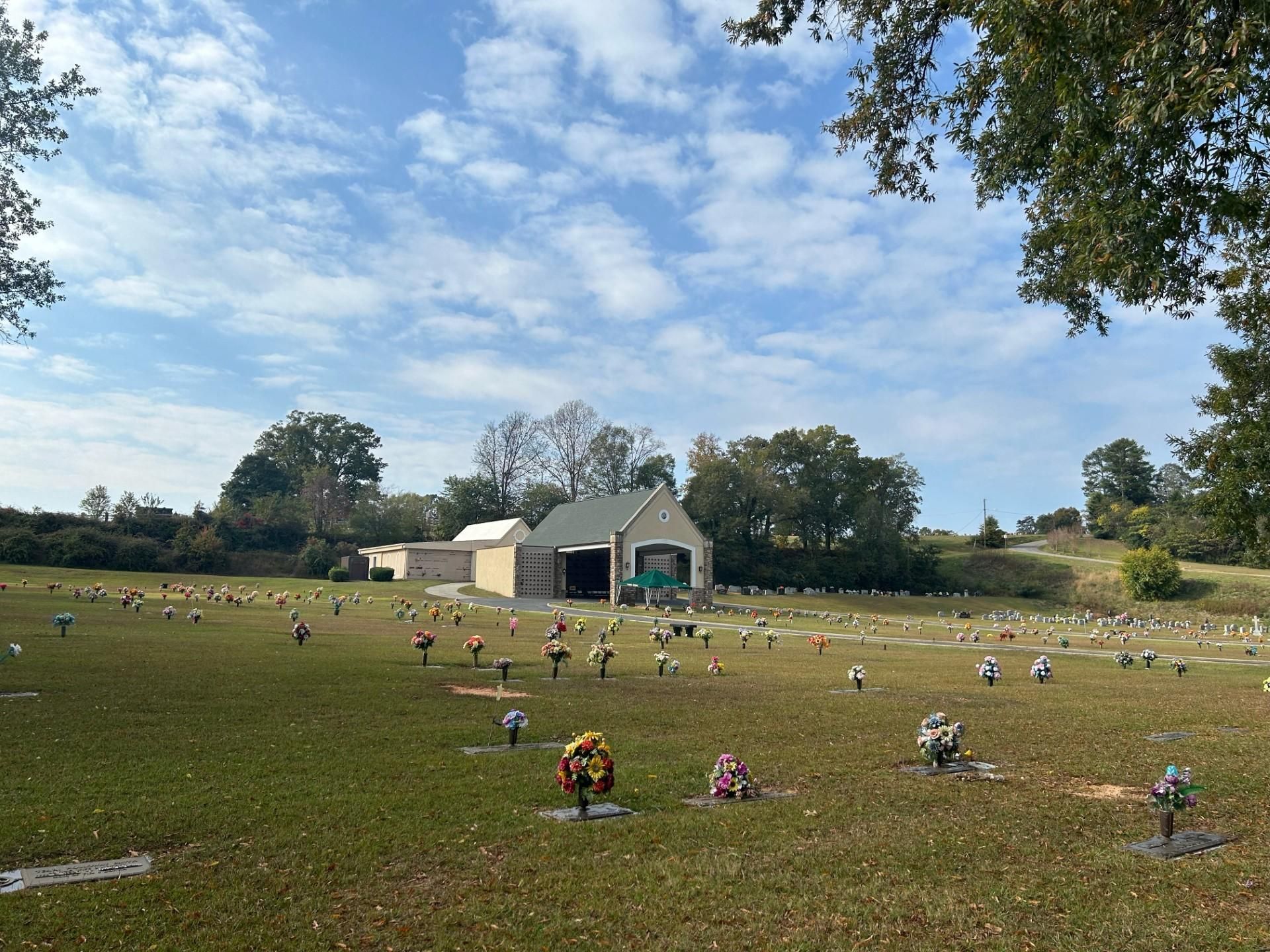 Cemetery with many headstones, small building in background, green grass, cloudy sky.