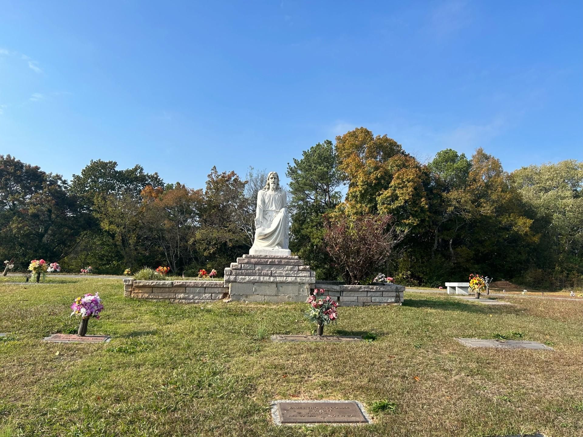 A statue of a robed figure stands in a cemetery with trees and flowers, under a blue sky.