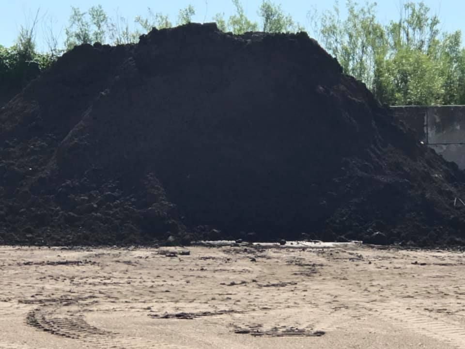 Large dark pile of soil or mulch in a dusty outdoor lot with trees in the background