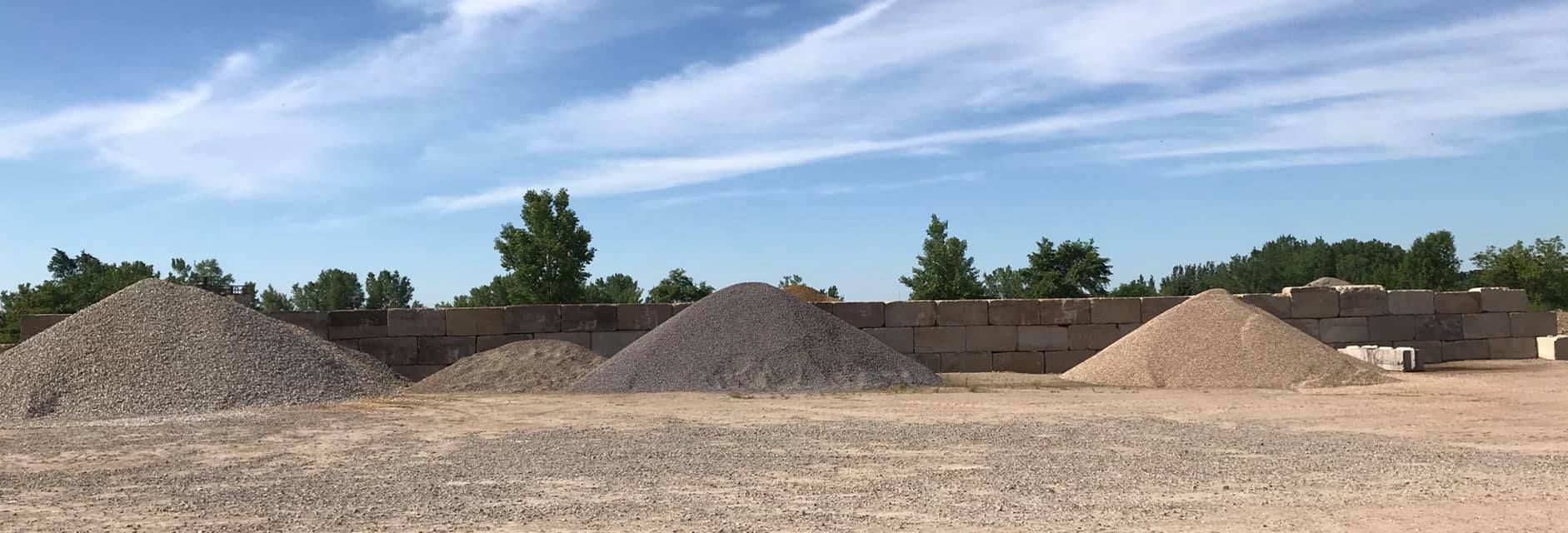 Gravel piles in a dirt lot with trees and blue sky in the background