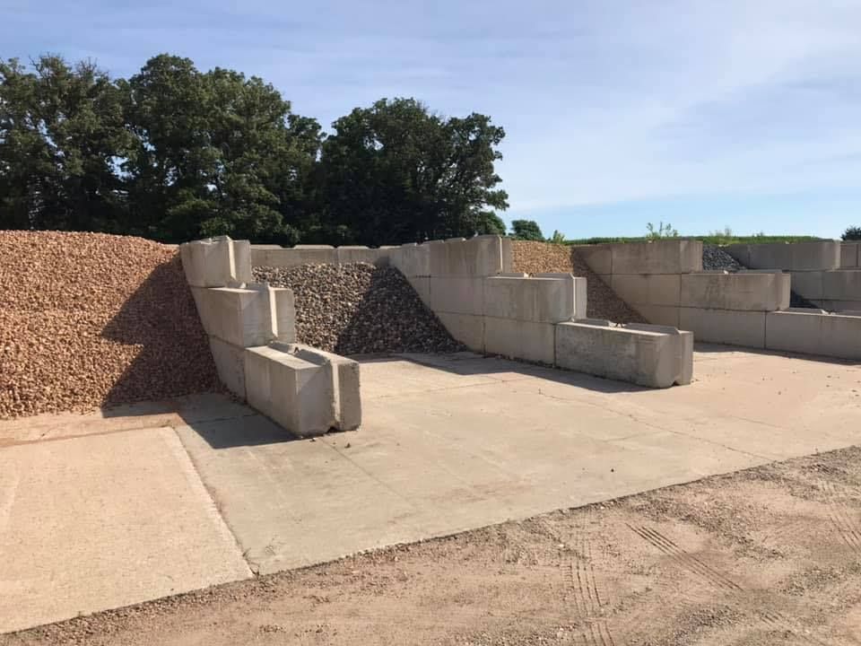 Concrete retaining wall blocks stacked in an outdoor yard beside gravel piles and trees