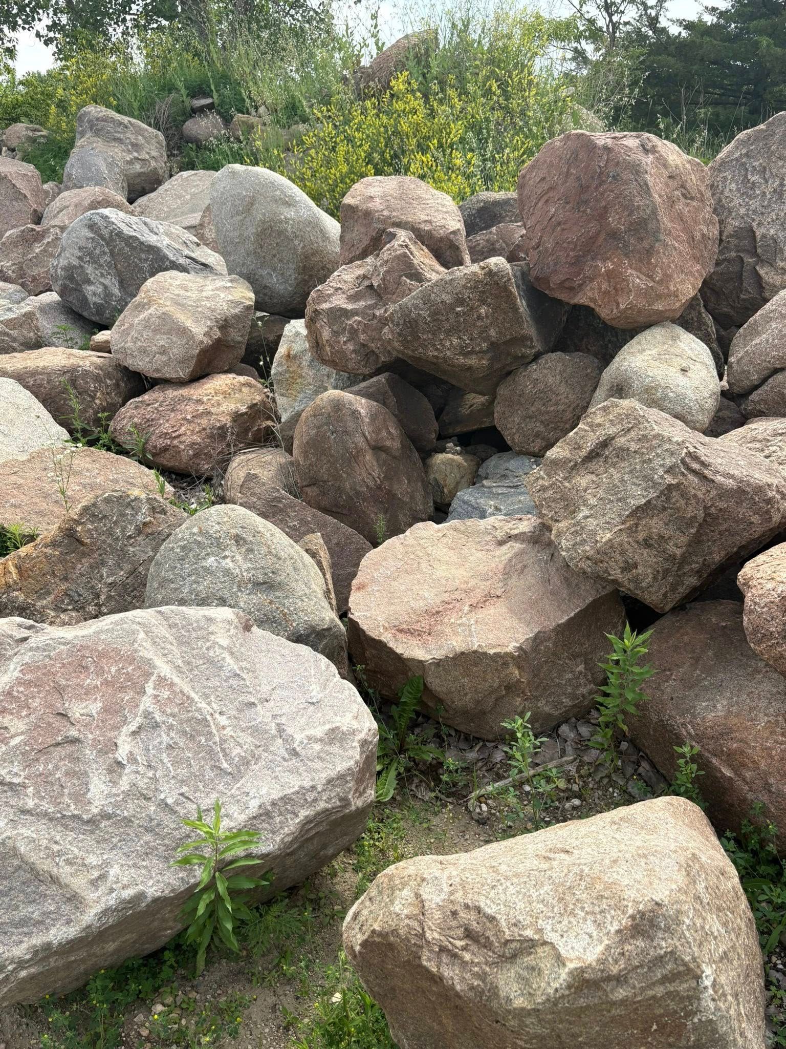 Pile of large gray and brown rocks stacked outdoors with green plants in the background