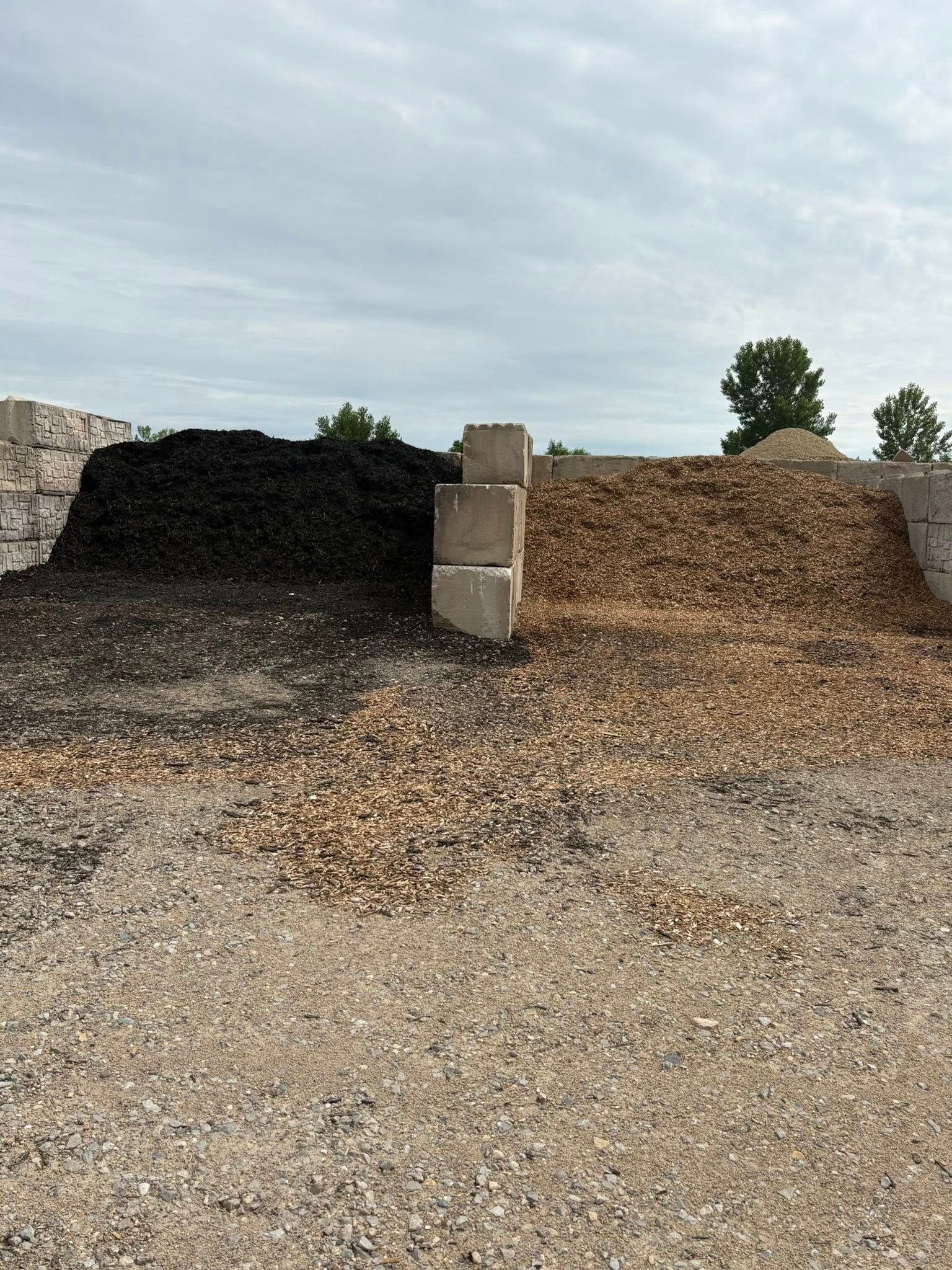 Piles of dark and tan gravel with a central concrete block in an outdoor yard under a cloudy sky
