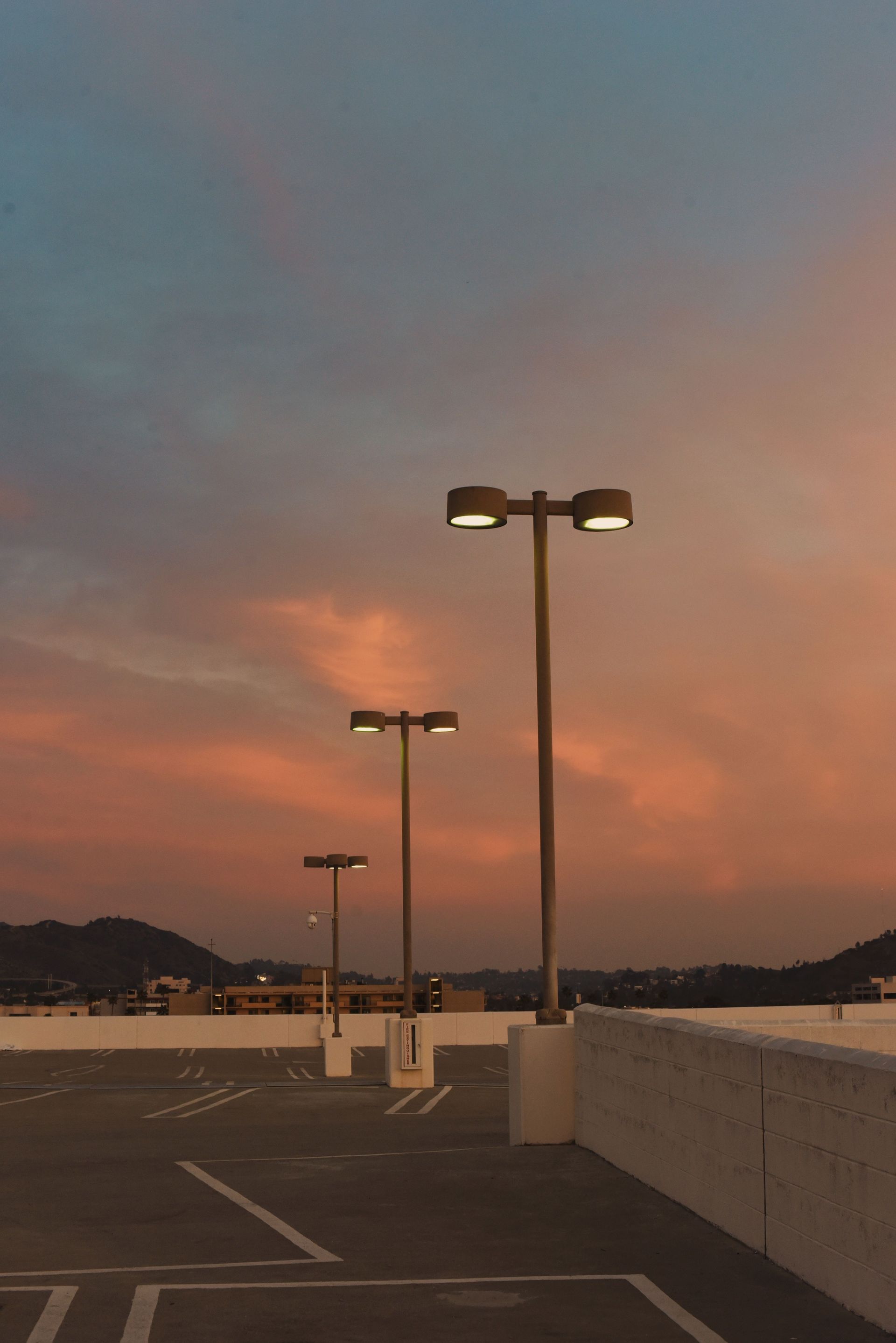 Parking lot with tall streetlights at dusk under a pink-orange sky over water and distant hills