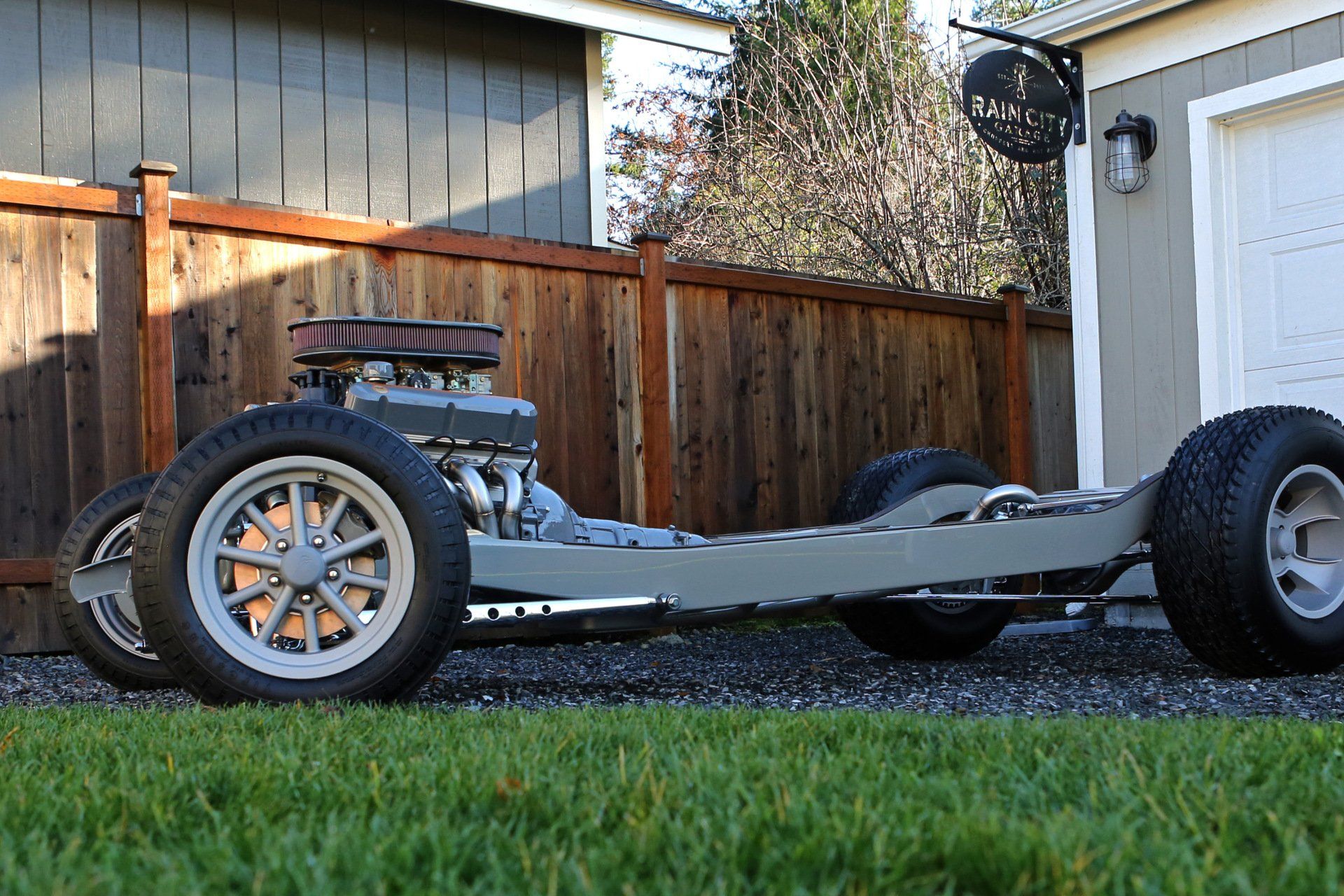 Low-slung custom car chassis with exposed engine and wheels parked in a driveway by a wooden fence