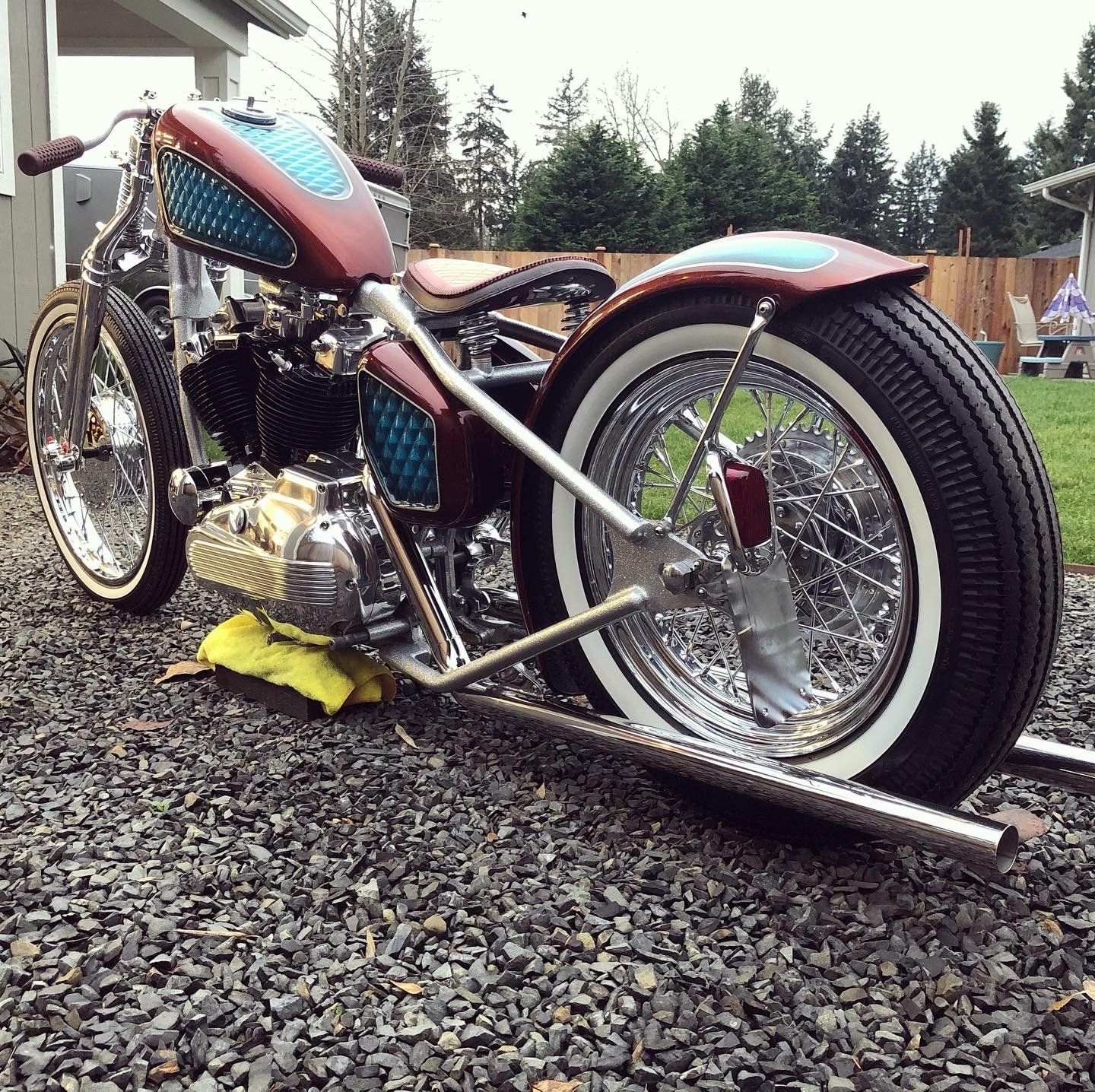 Custom red-and-blue lowrider motorcycle on gravel, side view with chrome details and whitewall tires.