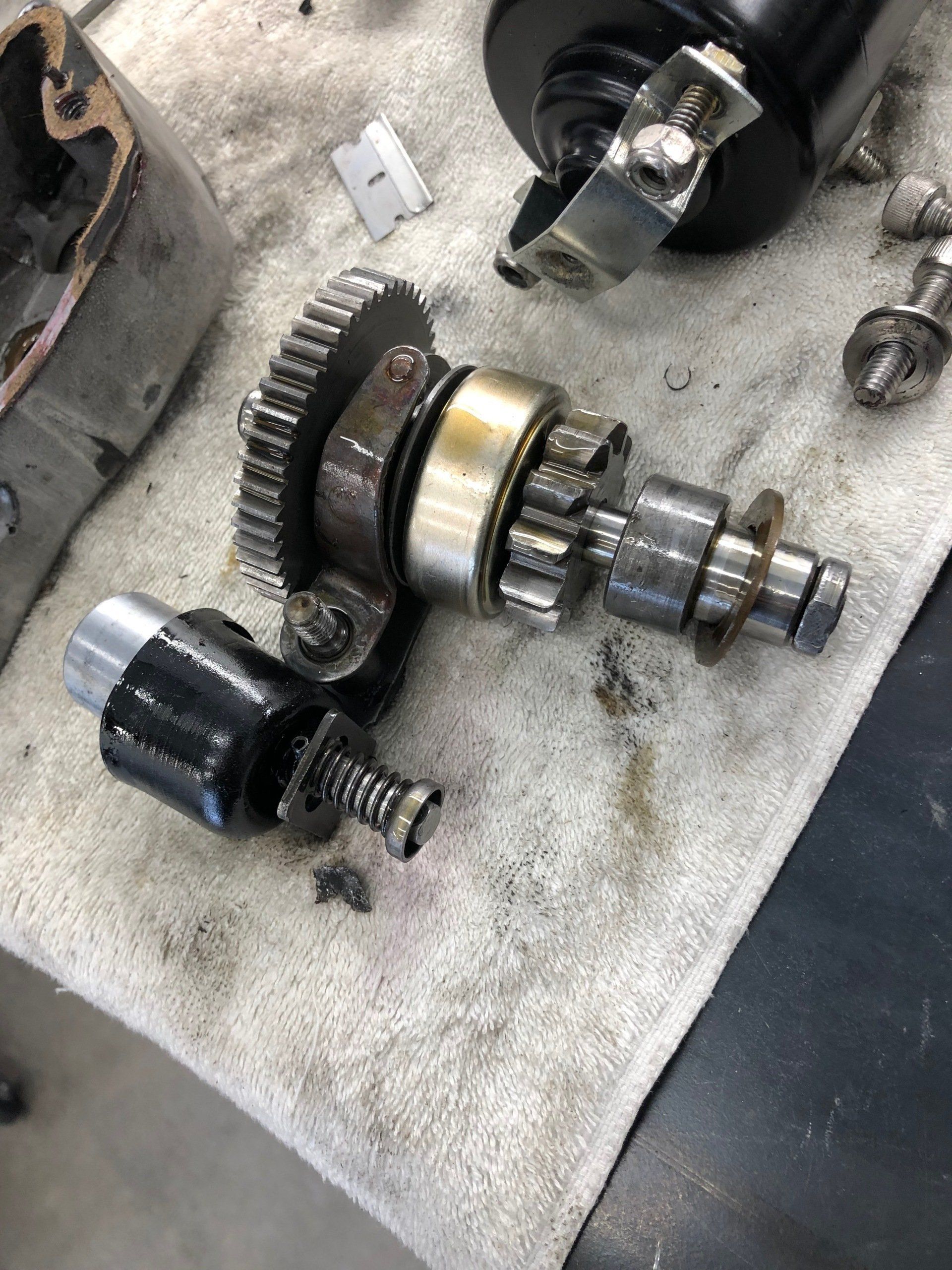 Assorted metal gears and mechanical parts on a workbench, including a gear shaft assembly and black plastic component