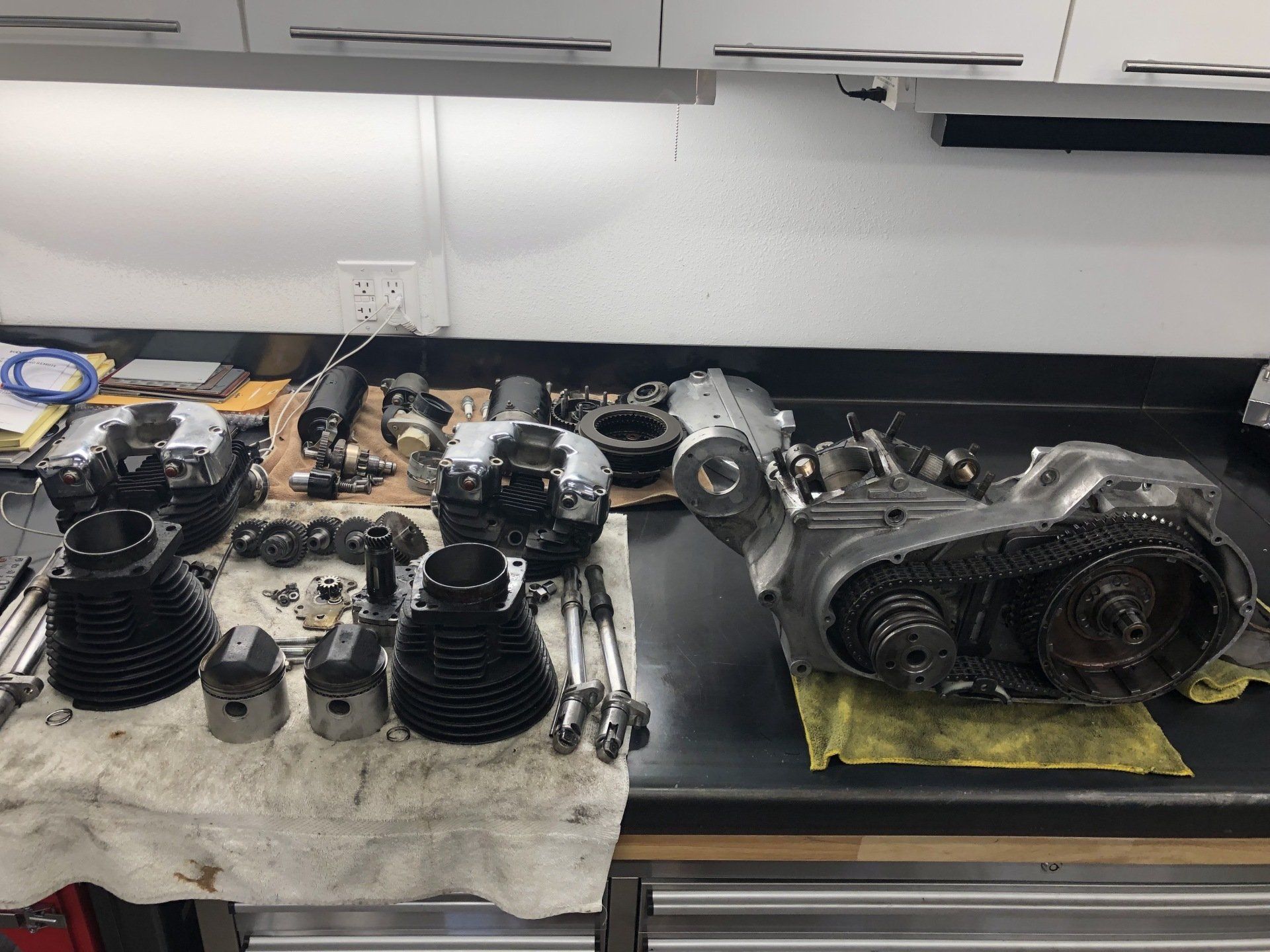 Engine parts laid out on a workshop bench under cabinets, including two black cylinder assemblies and a gearbox housing