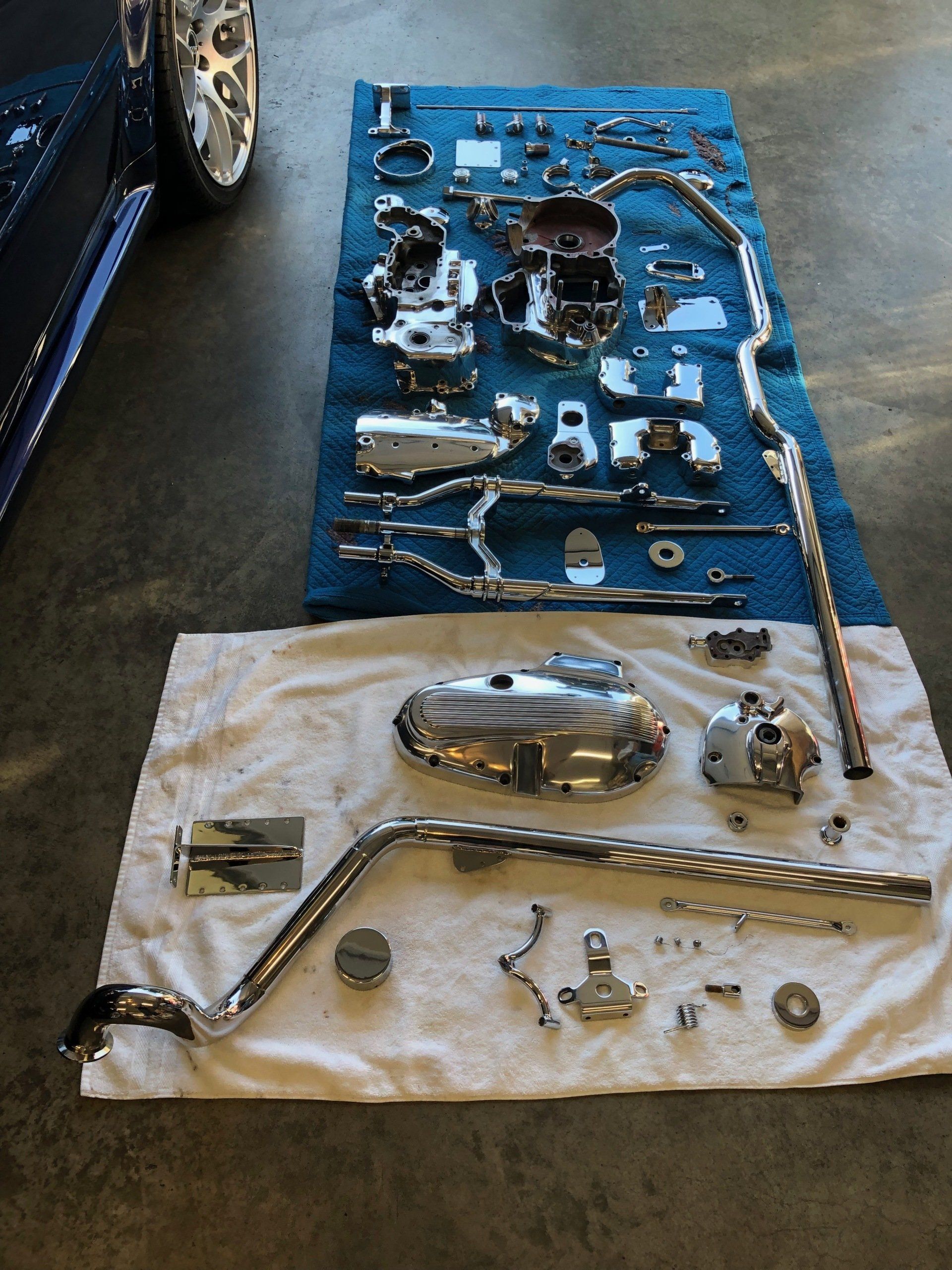 Car parts laid out on blue and white towels beside a car, including metal hoses and engine components
