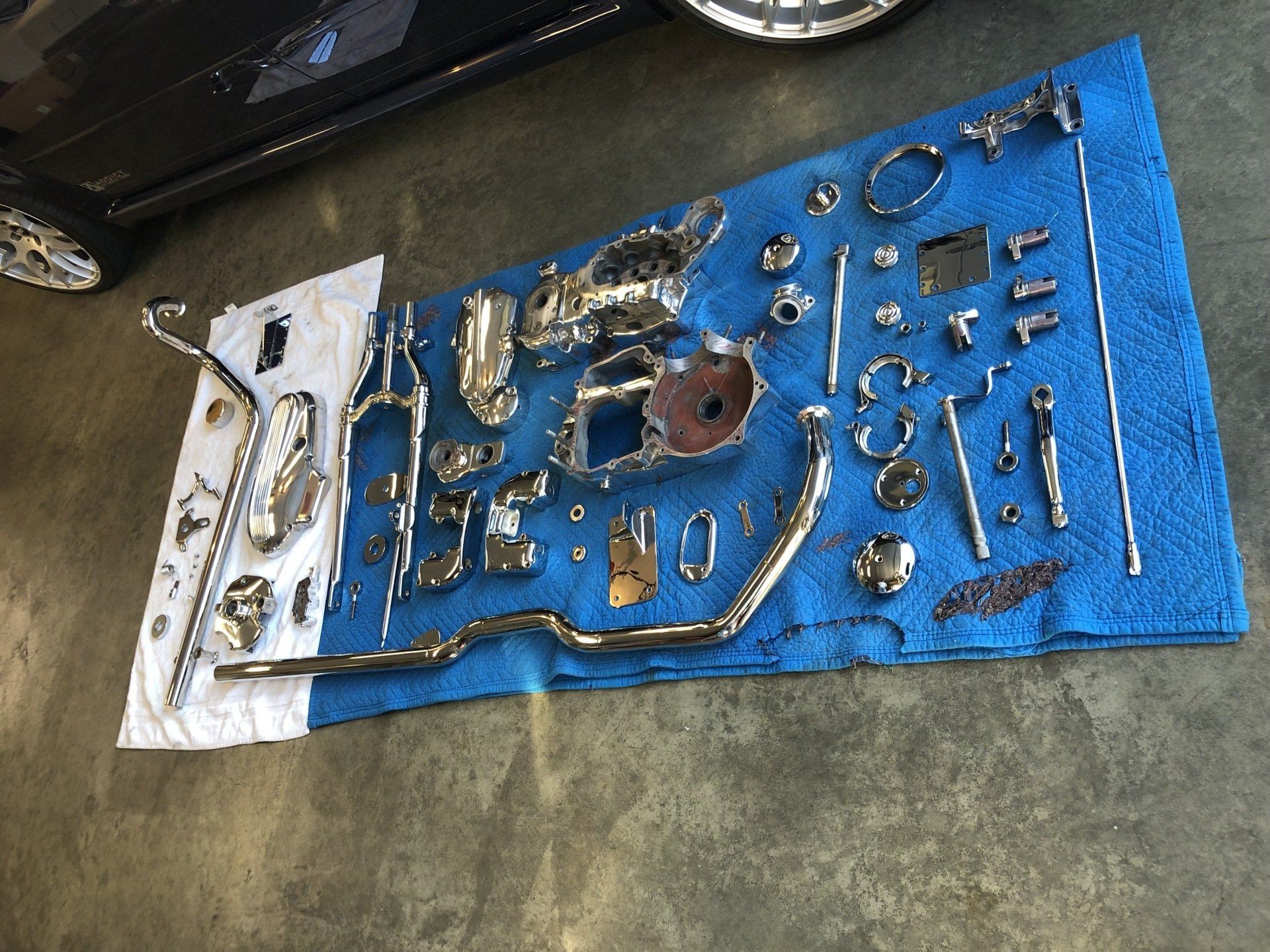 Car parts and tools laid out on a blue tarp on a concrete floor, with a car nearby.