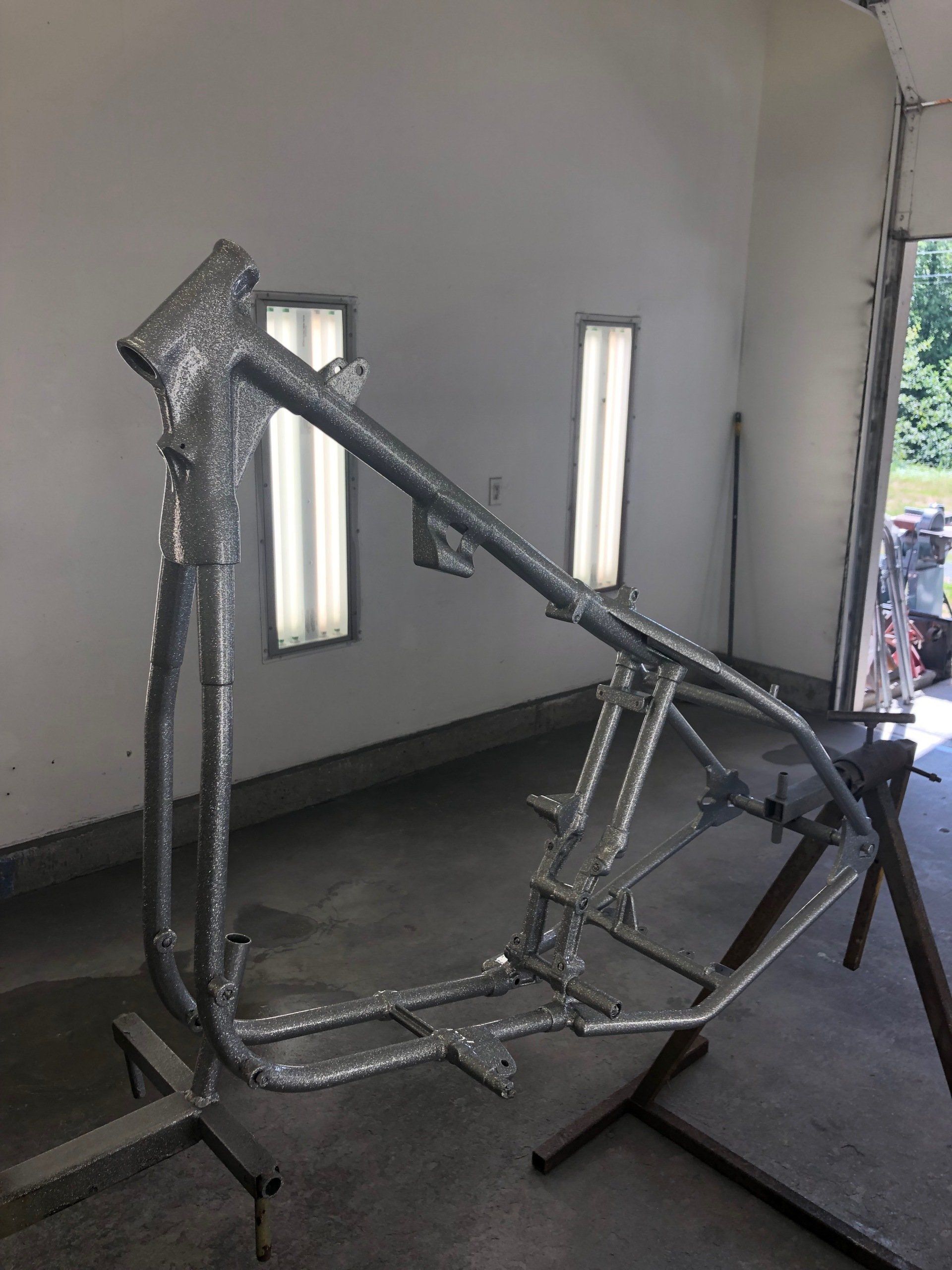 Metal tubular frame structure on stands in a workshop, shown unfinished and partially wrapped with tape.