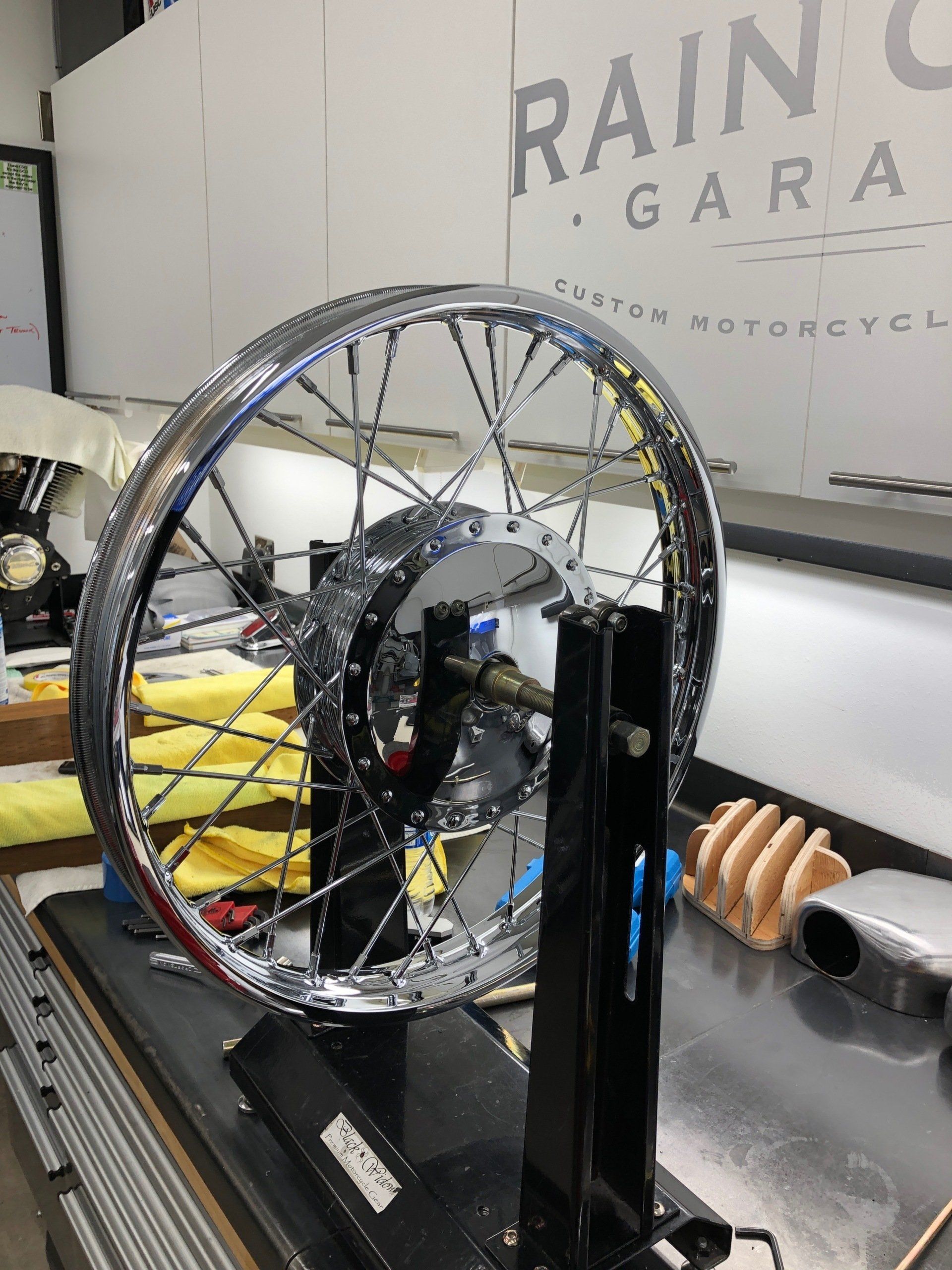 Chrome wheel mounted on a stand in a garage, with tools and a white vehicle in the background