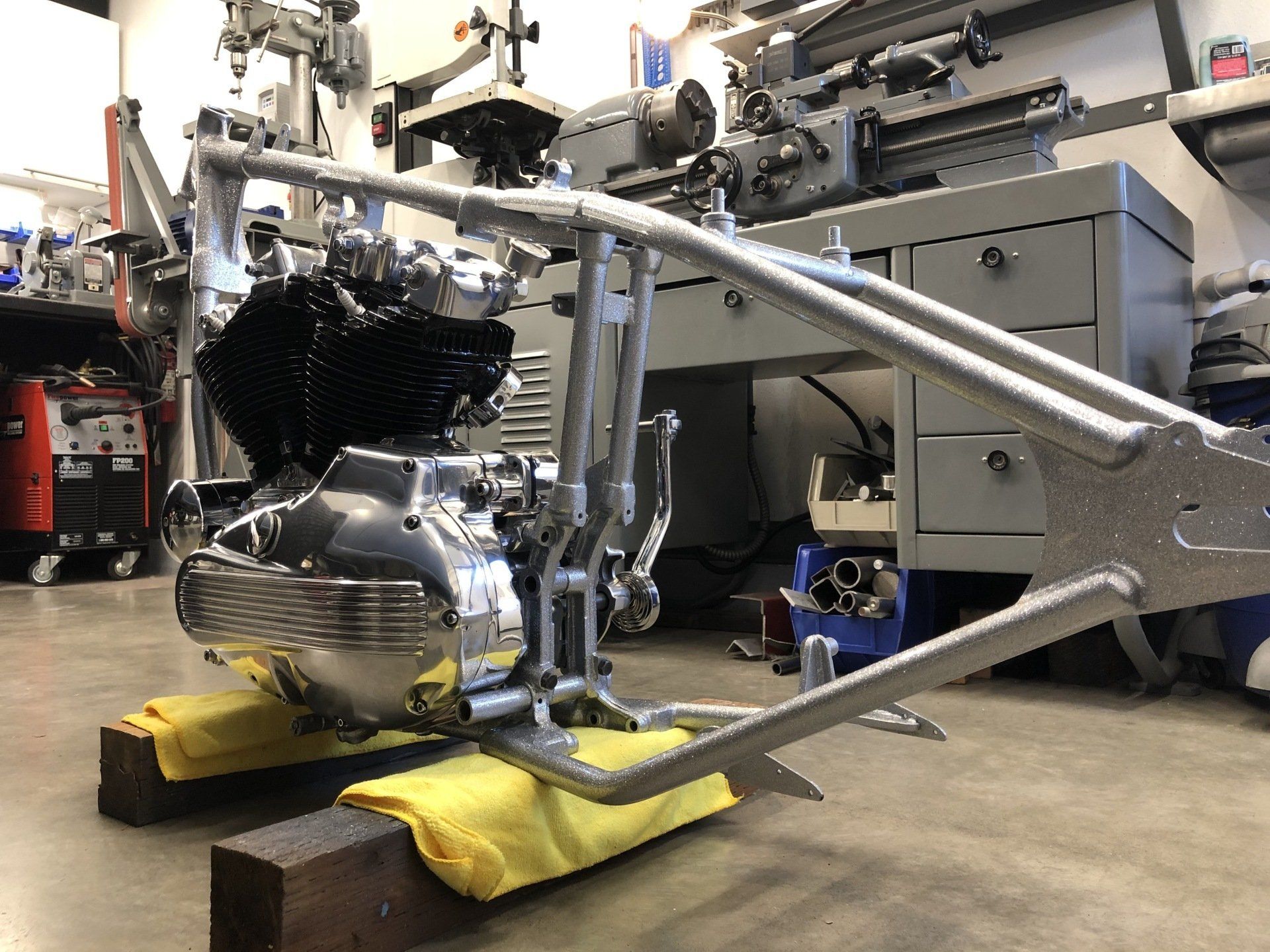 Motorcycle chassis on yellow stands in a workshop, showing engine and chrome frame under assembly