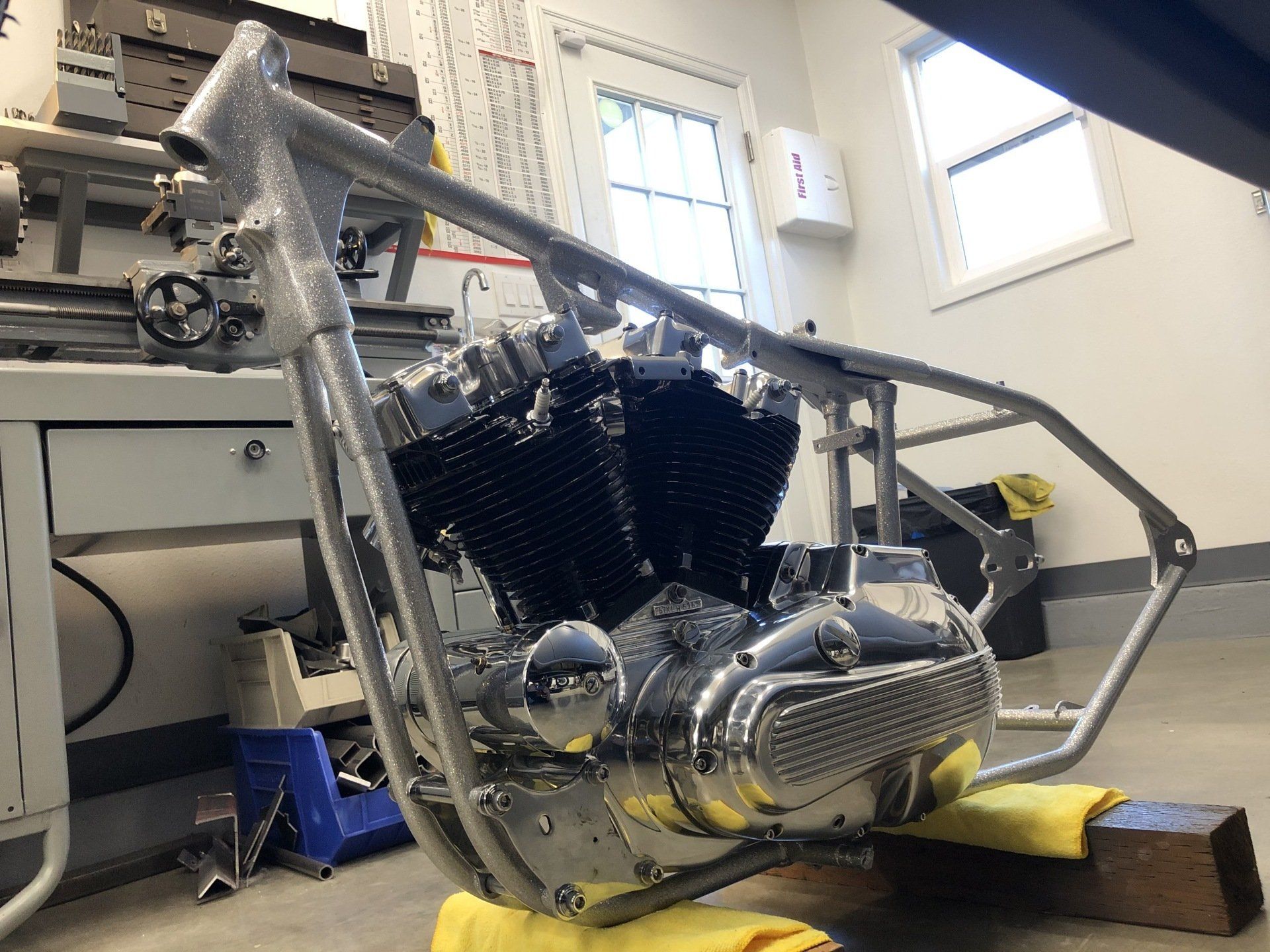 Custom motorcycle frame in a workshop, with chrome engine parts and a yellow wheel chock.