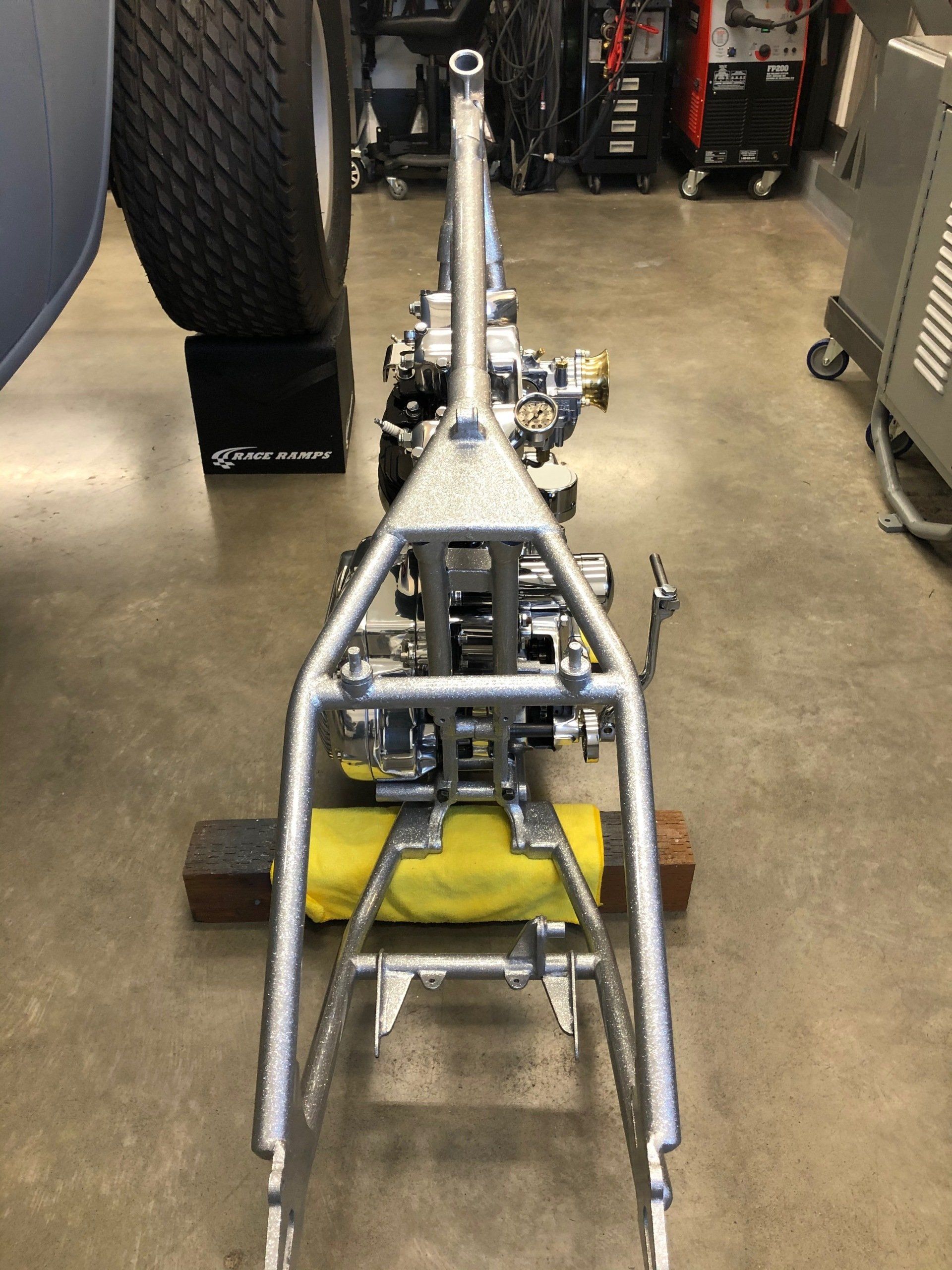 Front view of a small metal vehicle chassis in a workshop, with wheels and exposed components.