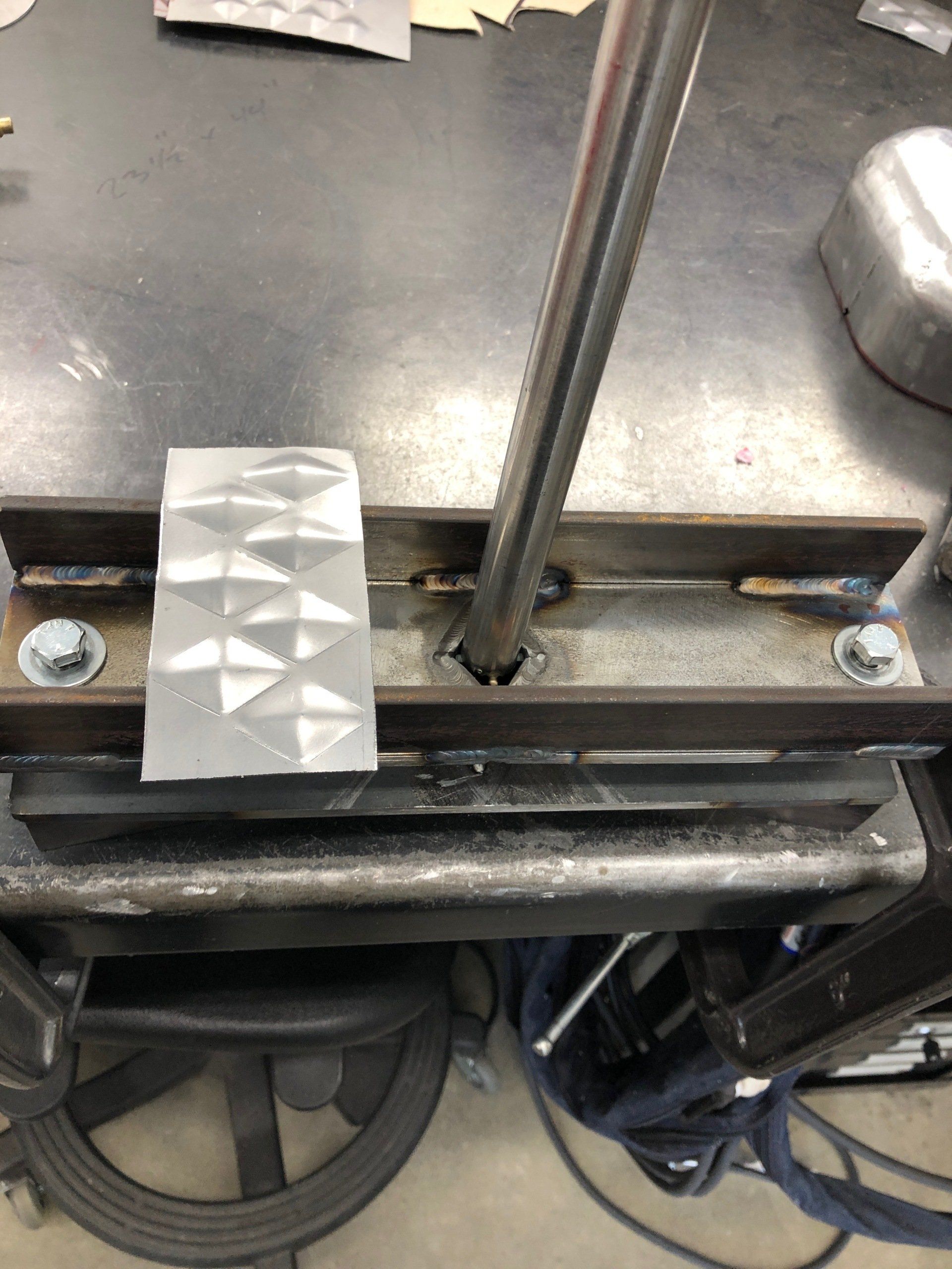 Close-up of a metal machine fixture with bolts, rods, and a textured silver block on a workbench.