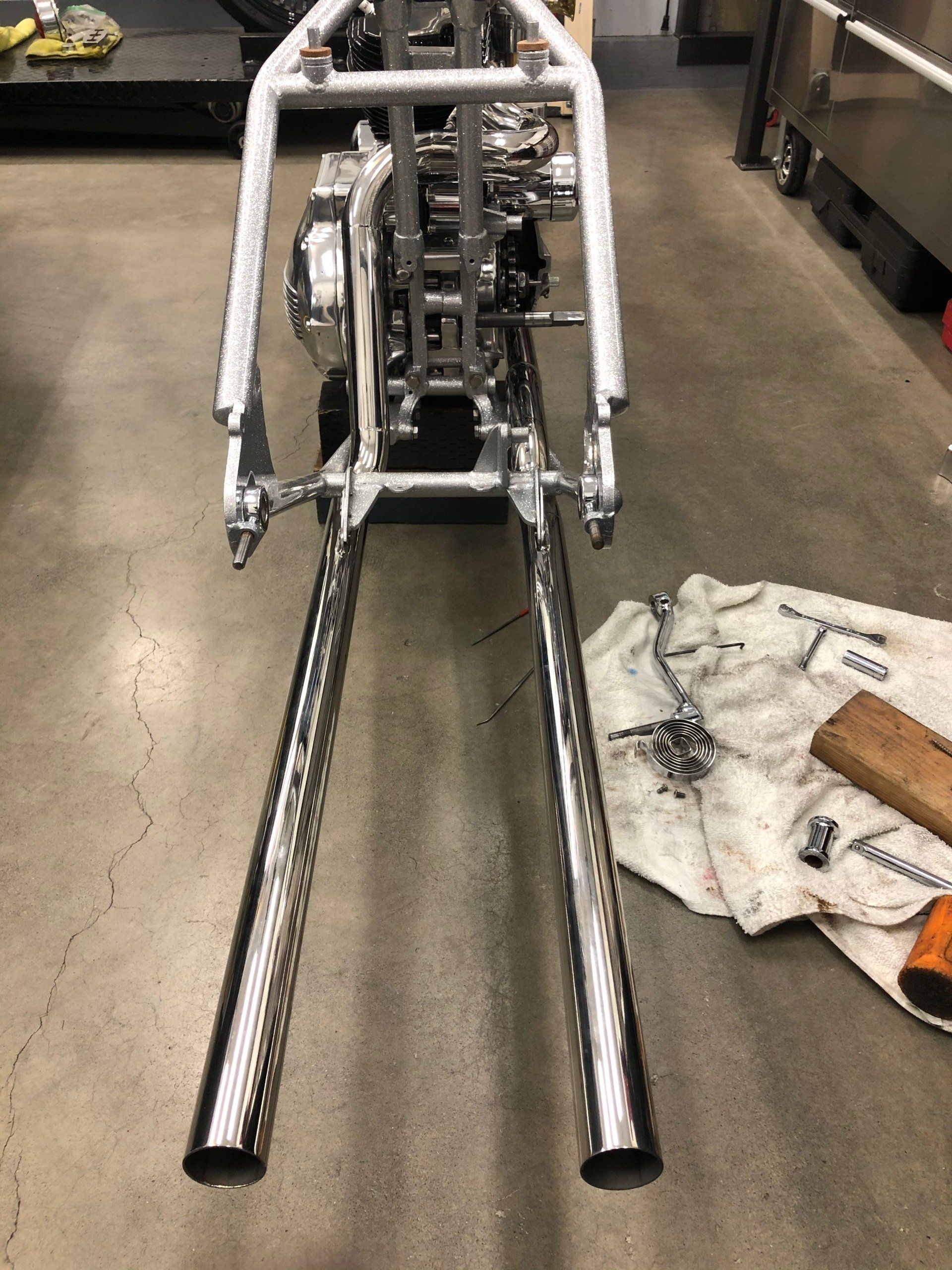 Motorcycle frame with long chrome fork tubes on a workshop floor, beside a white cloth and tools