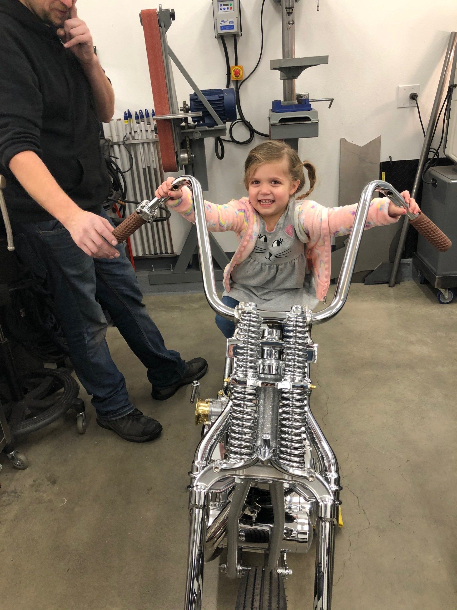 Child smiling on a chrome wheelchair prototype in a workshop, with an adult standing nearby.
