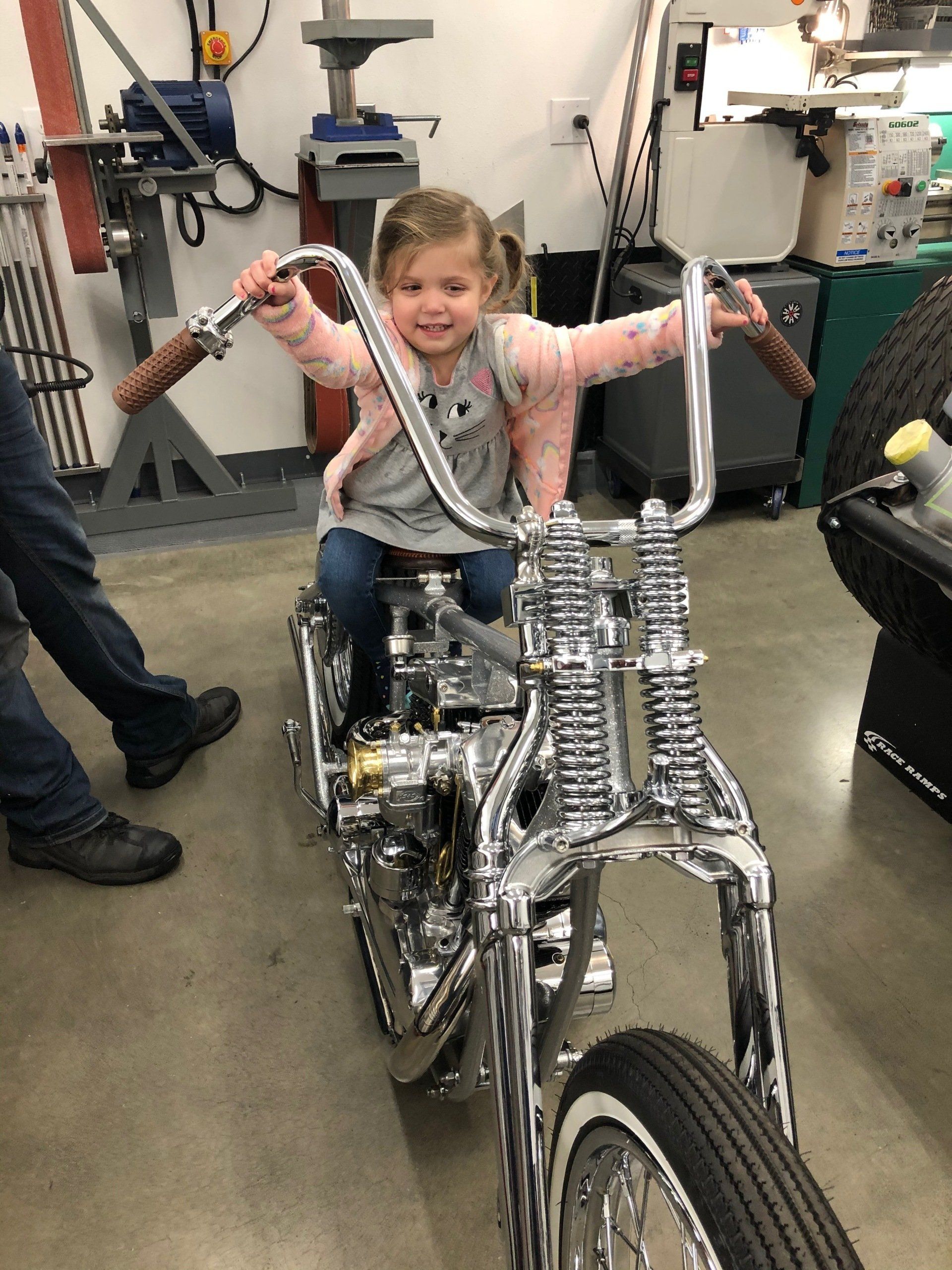 Child smiling on a silver bicycle sculpture in a workshop, arms outstretched with metal handlebars.