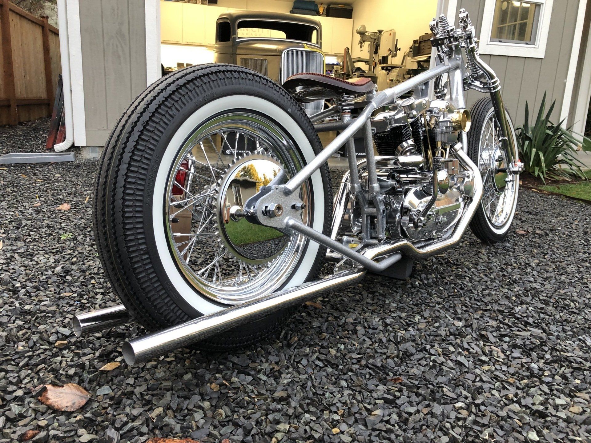 Custom chrome motorcycle parked on gravel beside a house, with a large rear wheel and polished frame.
