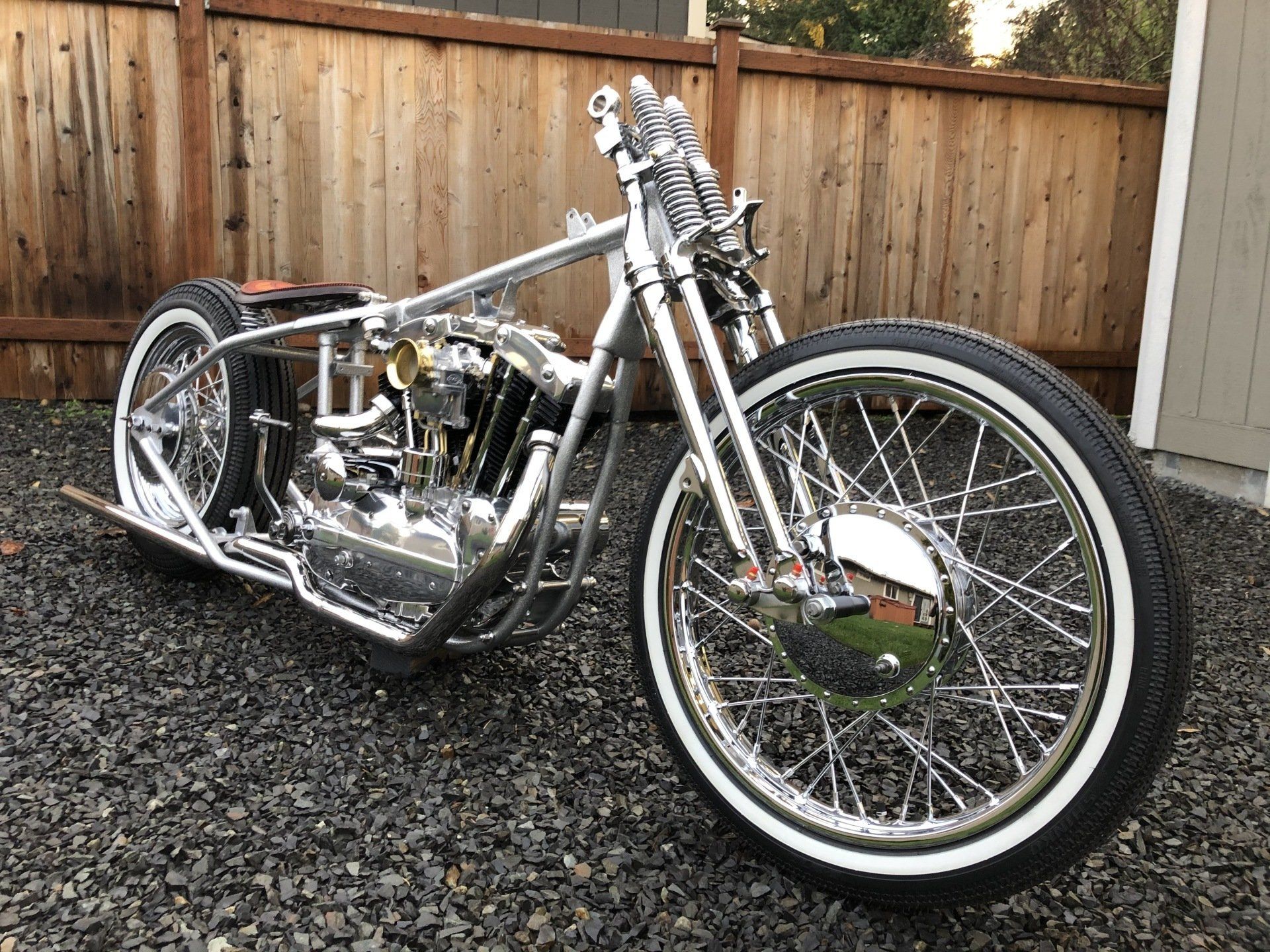 Custom silver chopper motorcycle parked on gravel in front of a wooden fence.