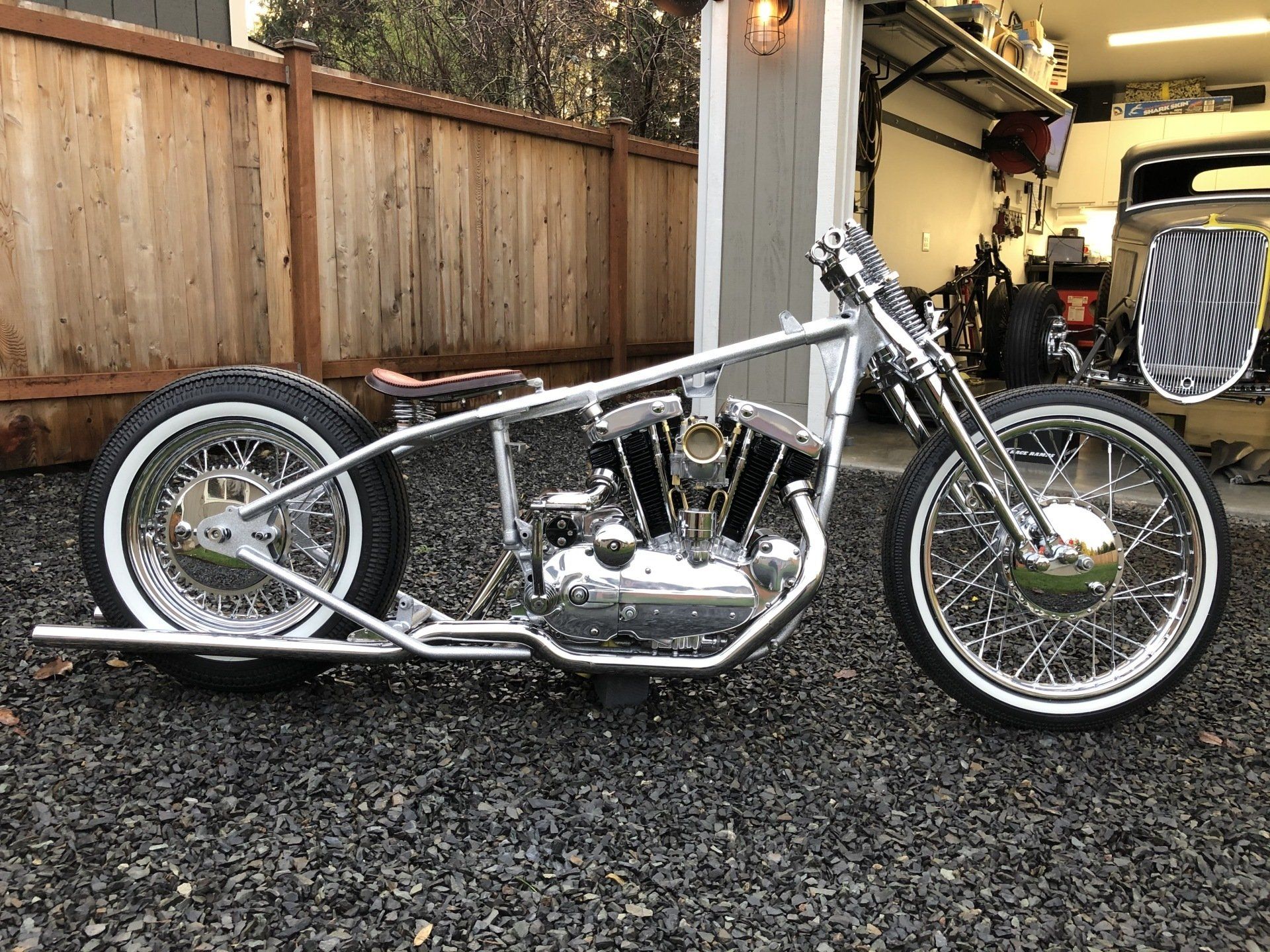 Custom silver chopper motorcycle parked on gravel in a driveway beside a wooden fence.