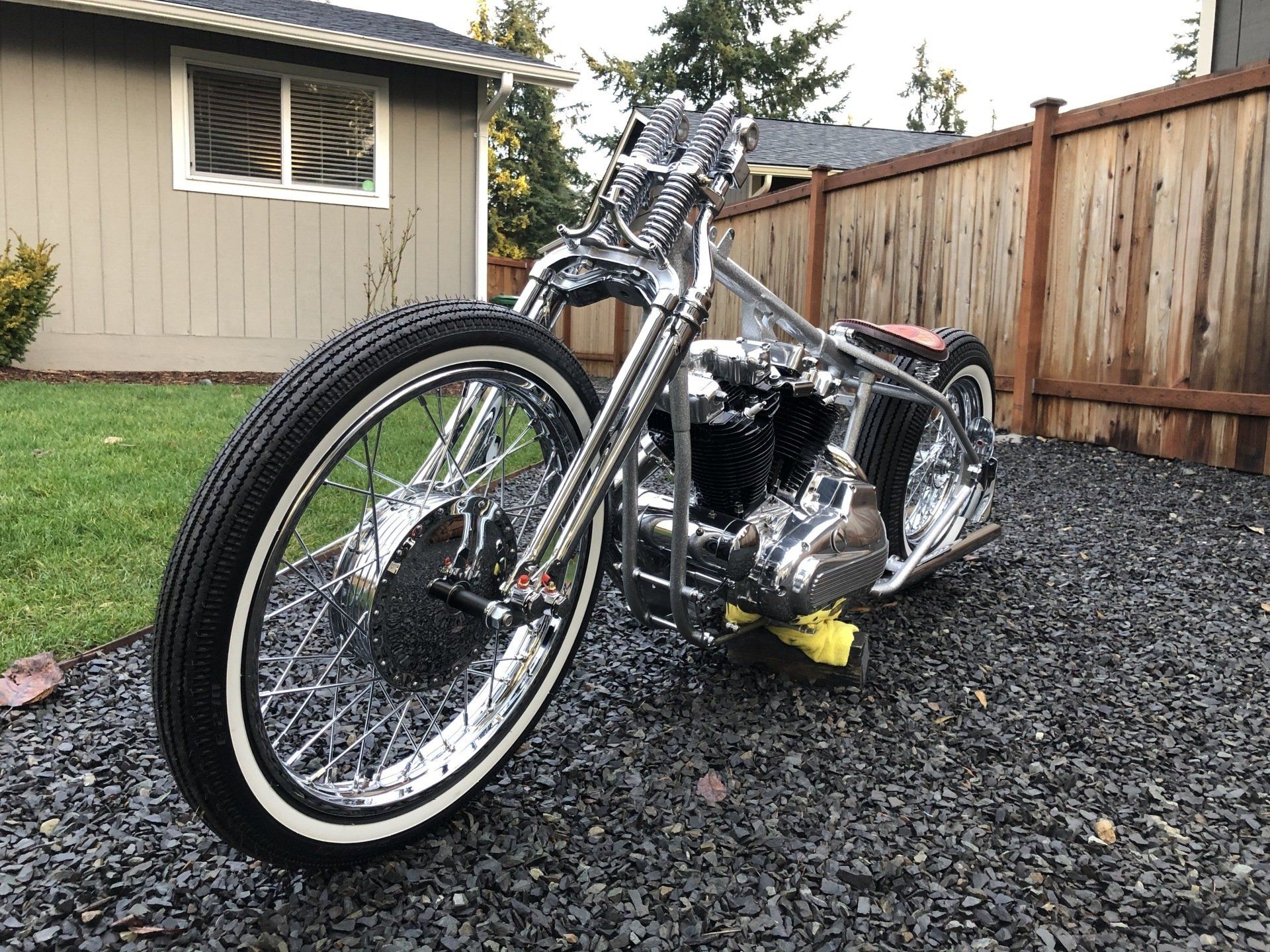 Chrome custom motorcycle on a gravel driveway beside a wooden fence and house