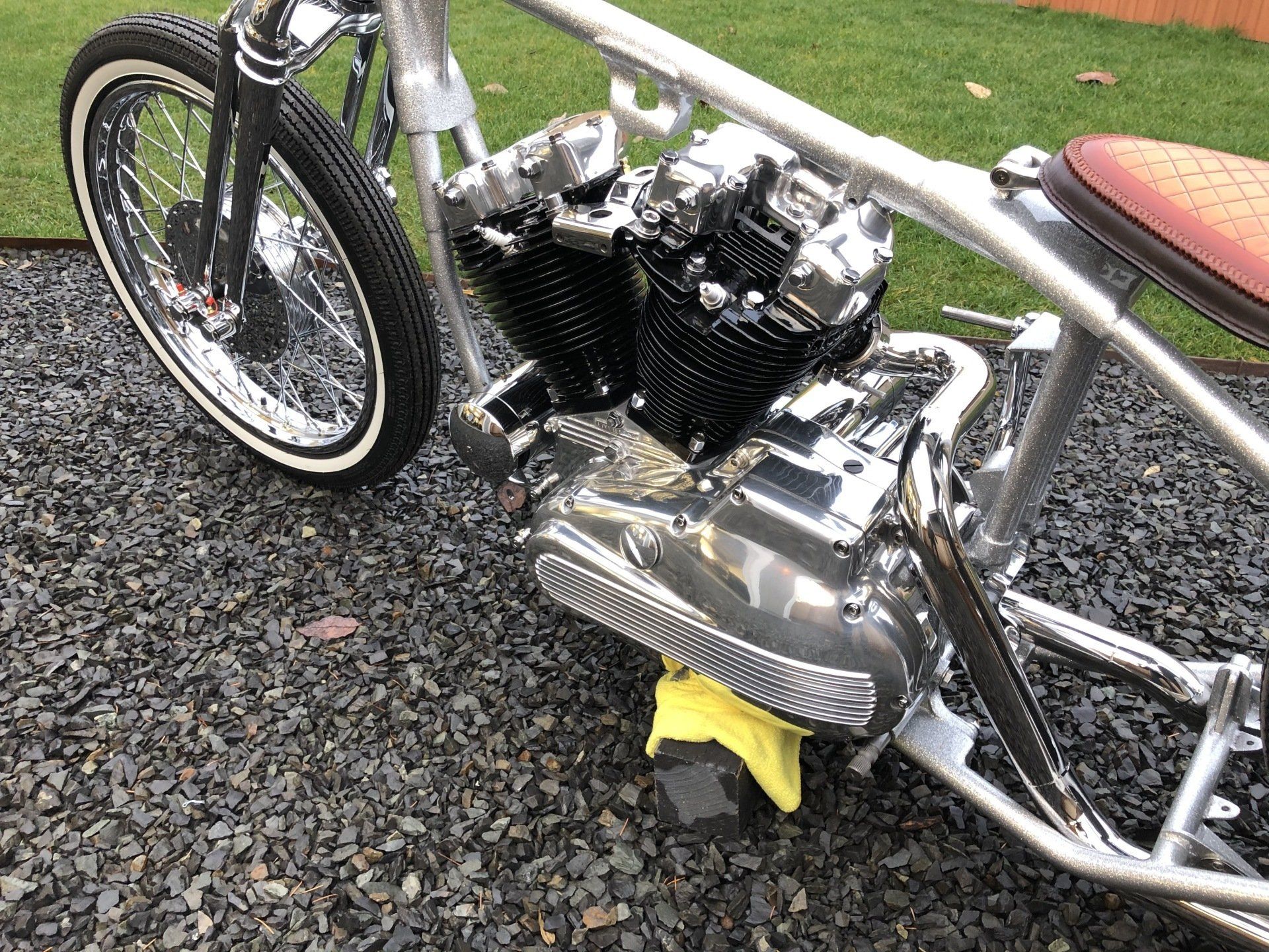 Custom chrome motorcycle parked on gravel beside grass, with a bright yellow cloth under the engine.