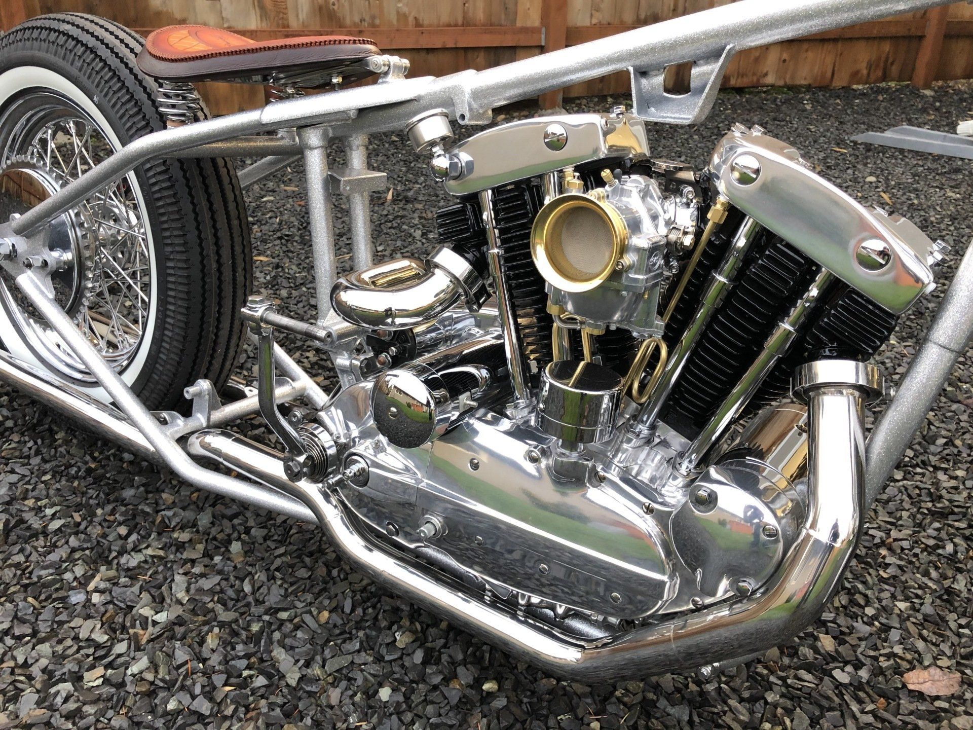 Chrome custom motorcycle engine and frame on gravel, with a wooden fence in the background