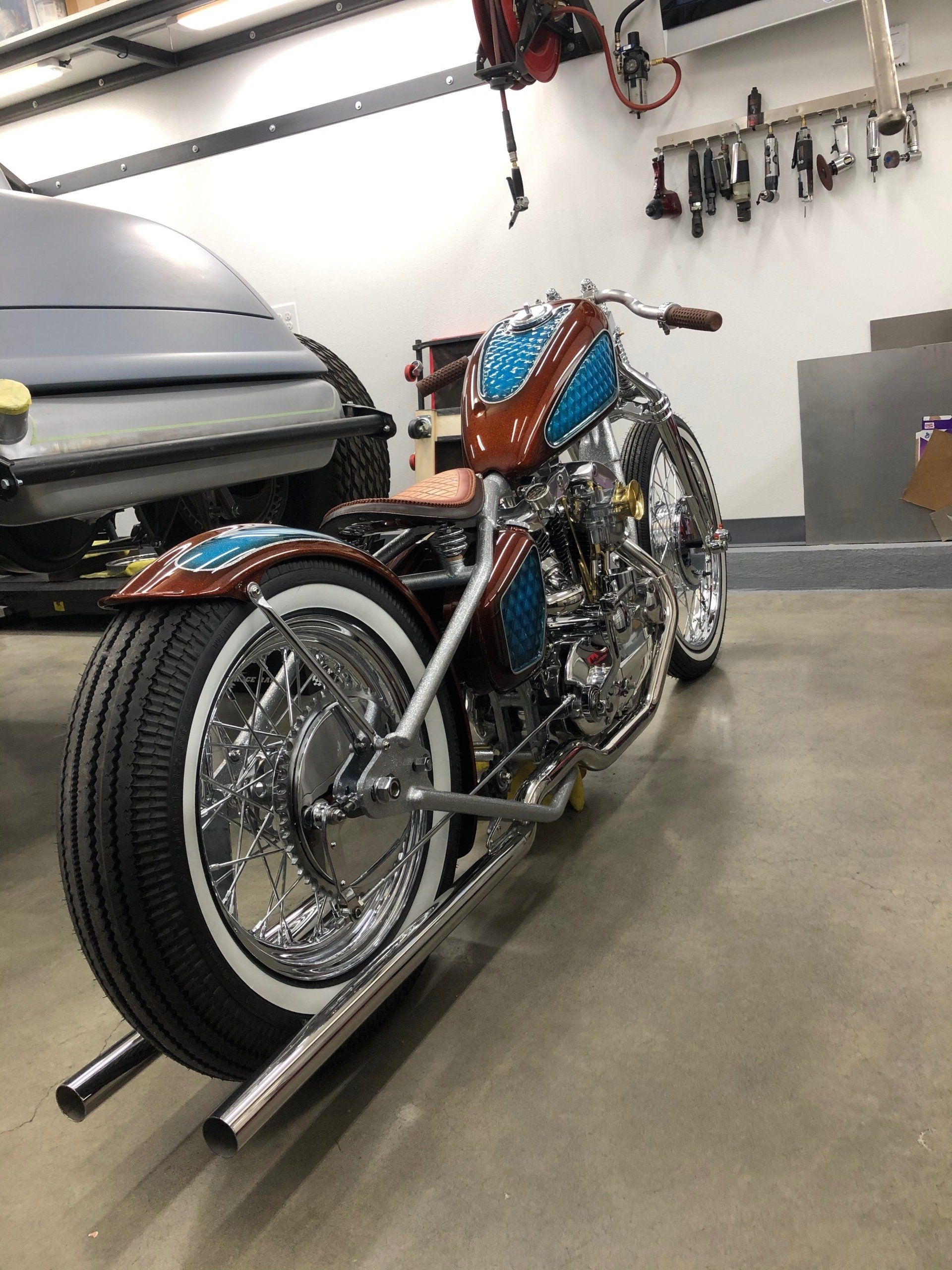 Custom blue-and-orange chopper motorcycle in a garage, shown from the rear-left side.