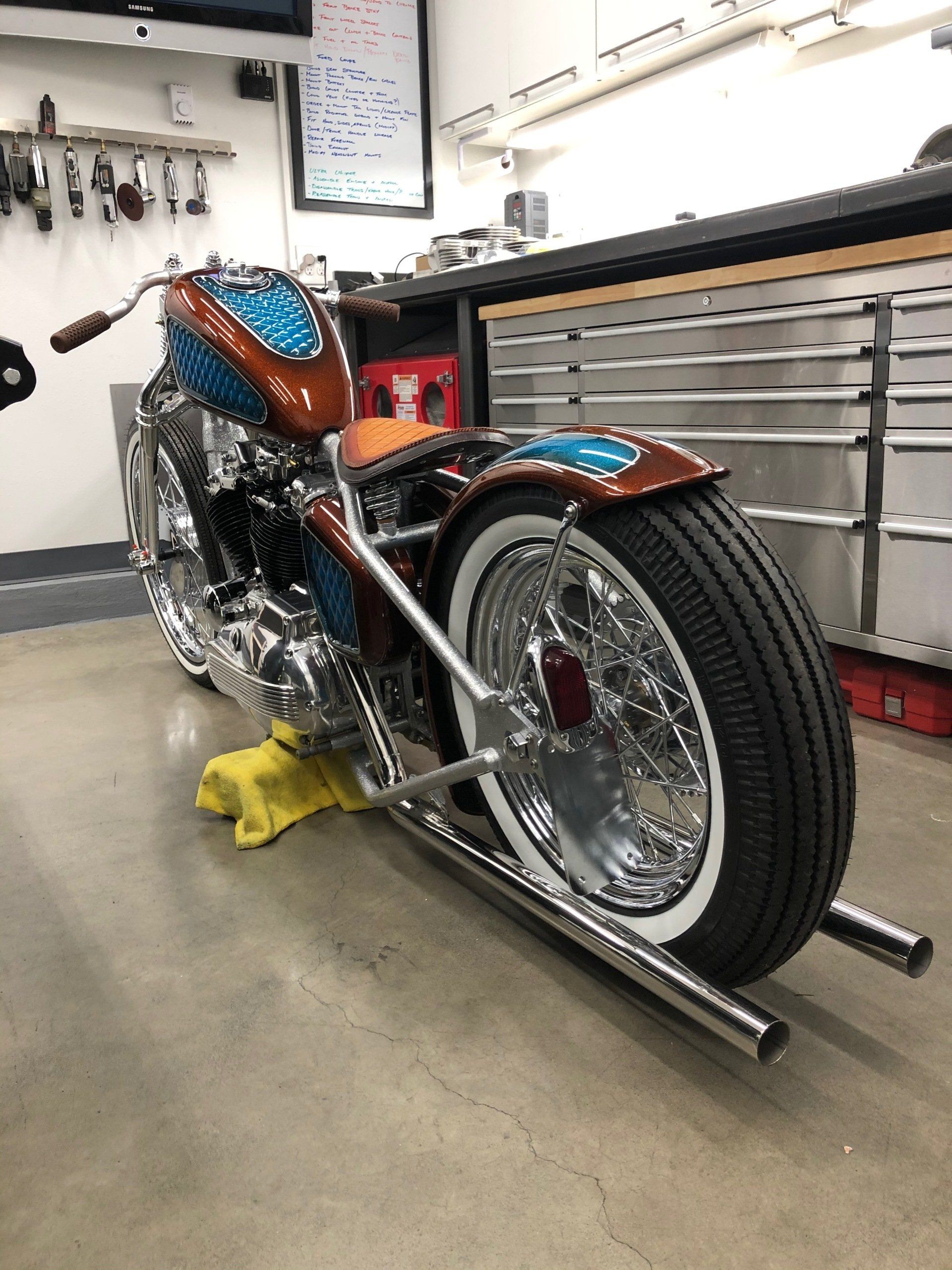 Custom blue-and-orange motorcycle on a stand in a workshop, with chrome details and a front wheel chock.