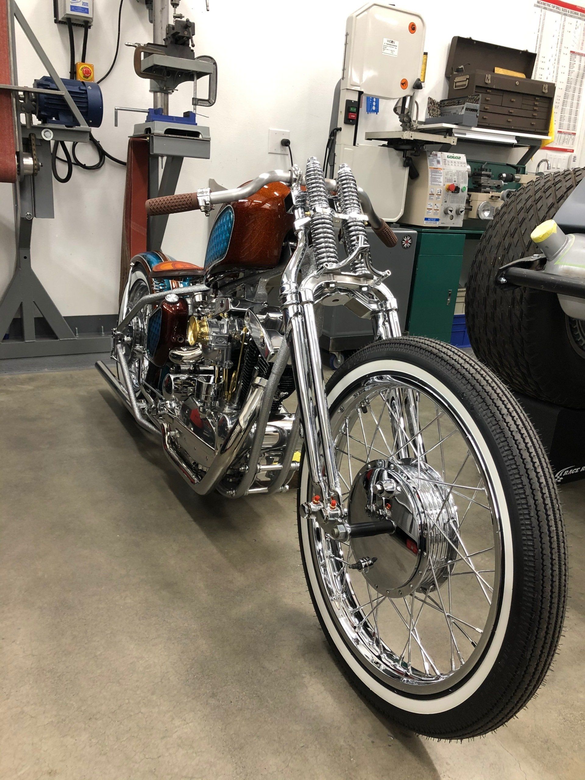 Custom chrome motorcycle in a workshop, with whitewall front tire and orange gas tank.