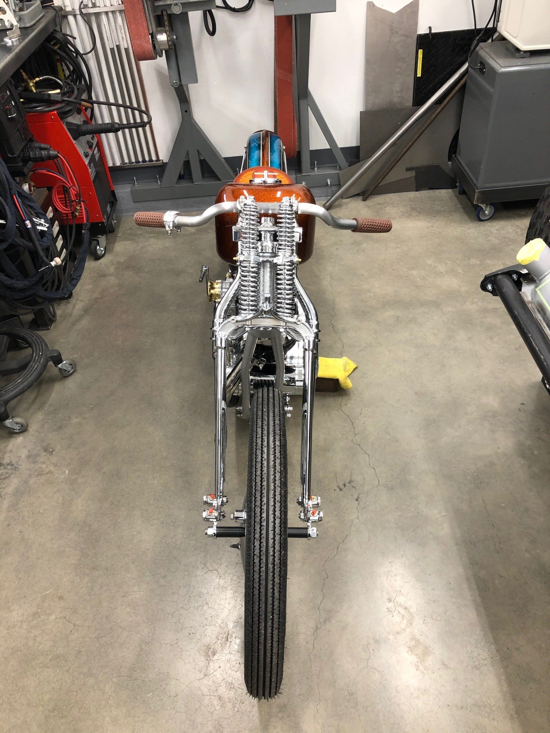 Custom motorcycle frame in a workshop, viewed from the front with handlebars and front wheel visible.