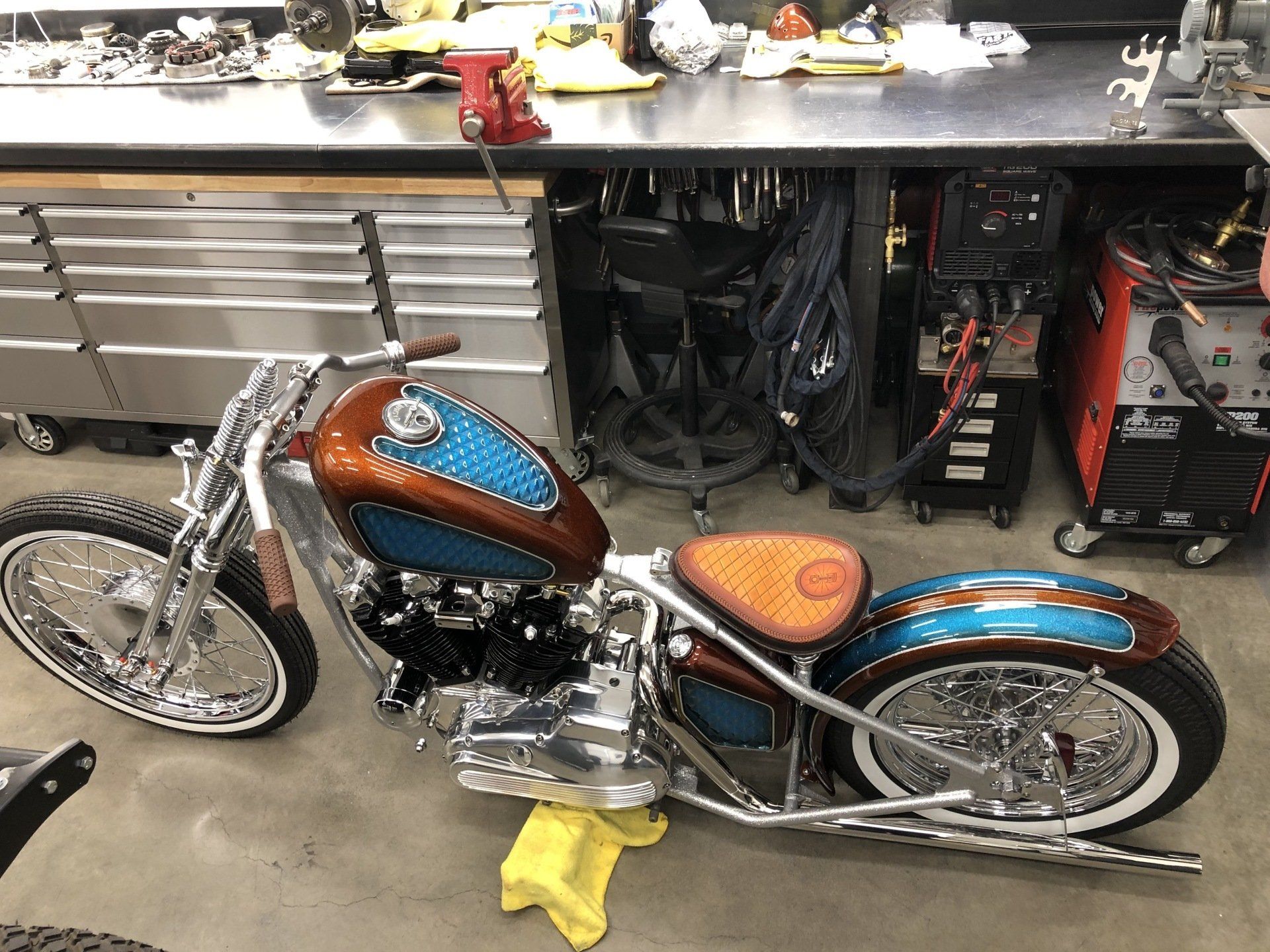 Custom blue-and-brown chopper motorcycle displayed in a garage workshop beside tool cabinets and equipment