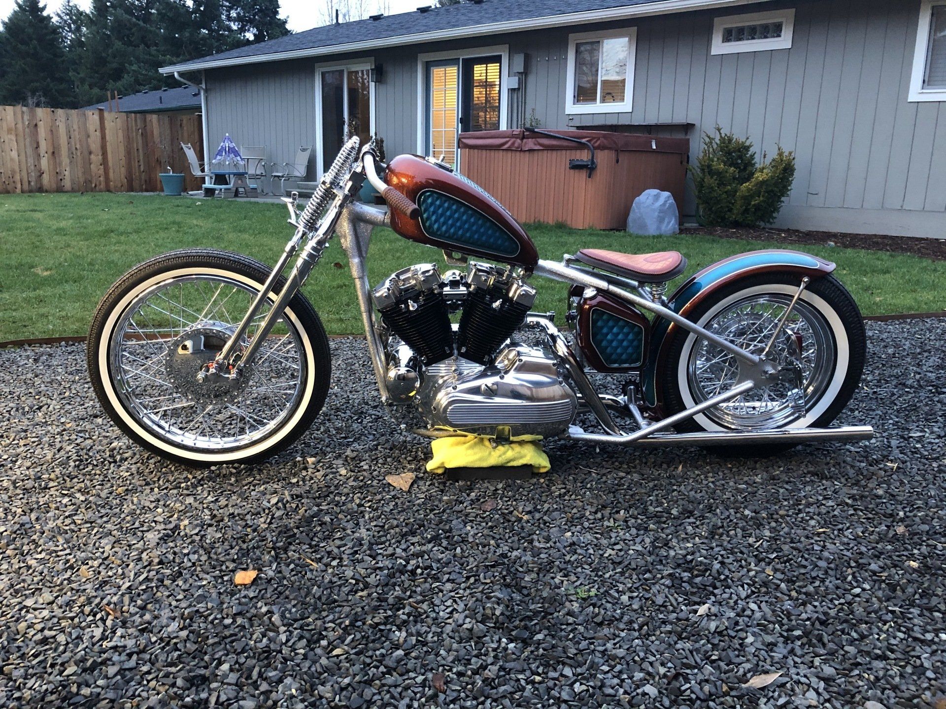 Custom chopper motorcycle with orange tank parked on gravel beside a house