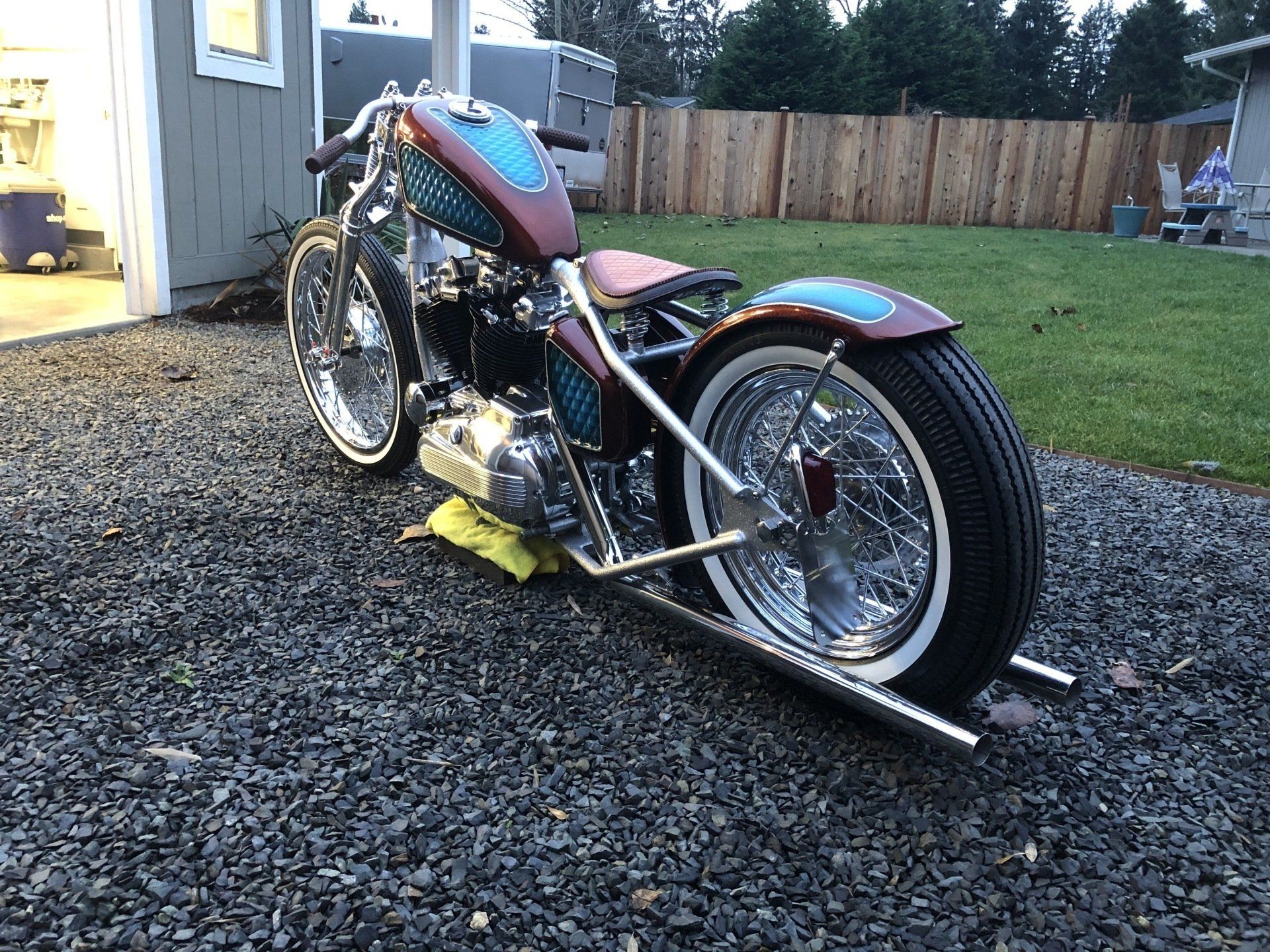 Custom chopper motorcycle parked on gravel beside a shed, with a wooden fence and lawn in the background