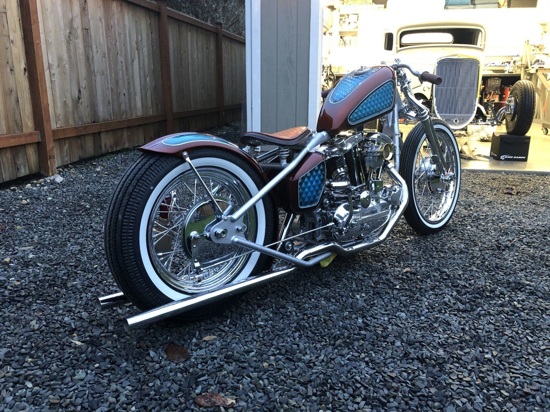 Custom chopper motorcycle parked on gravel beside a wooden fence, with garage in the background