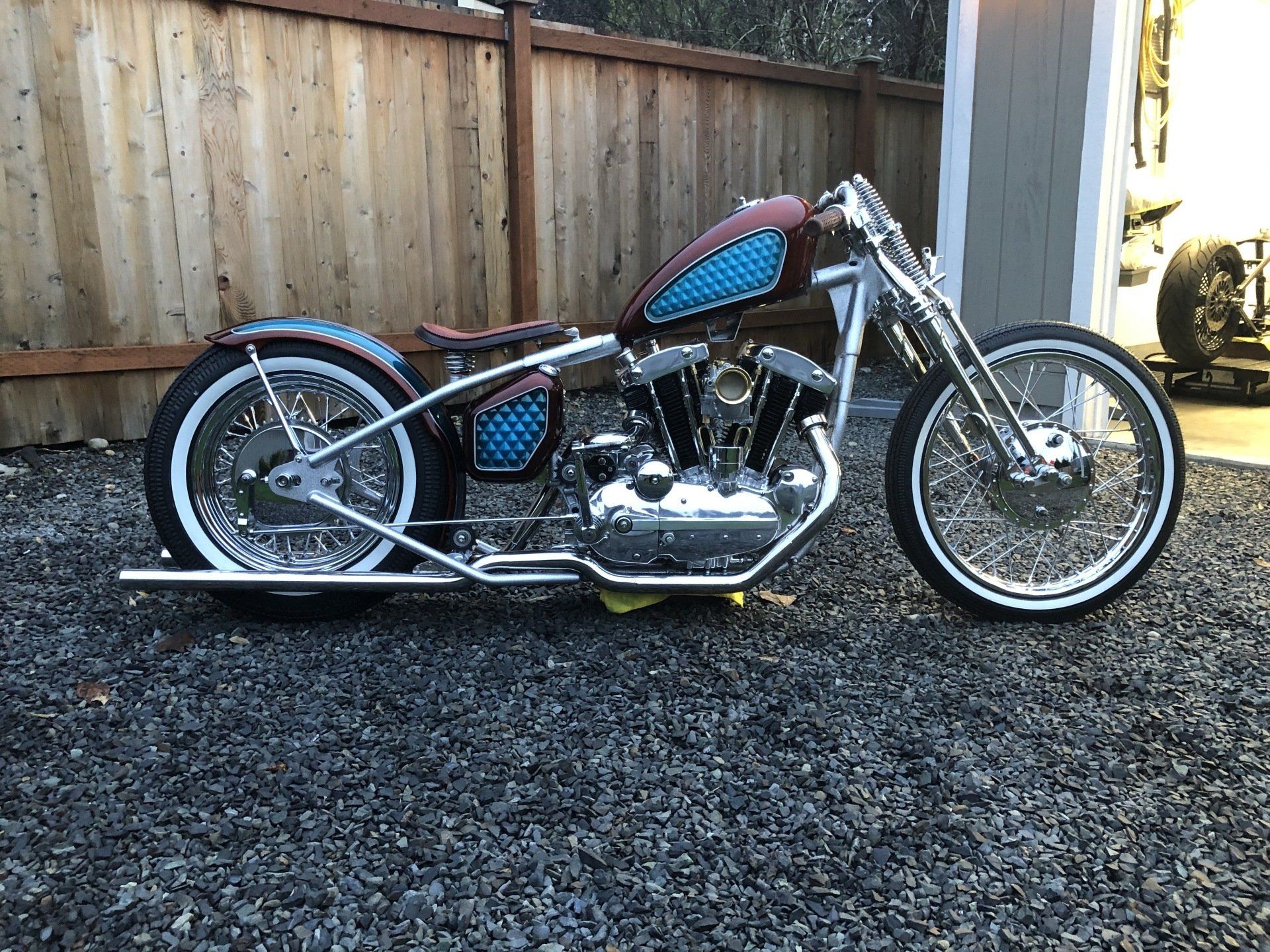 Custom chopper-style motorcycle with blue and maroon tank parked on gravel beside a wooden fence