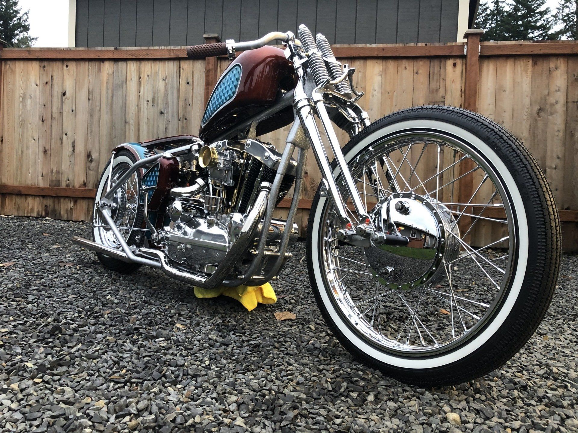 Custom chrome chopper motorcycle parked on gravel in front of a wooden fence
