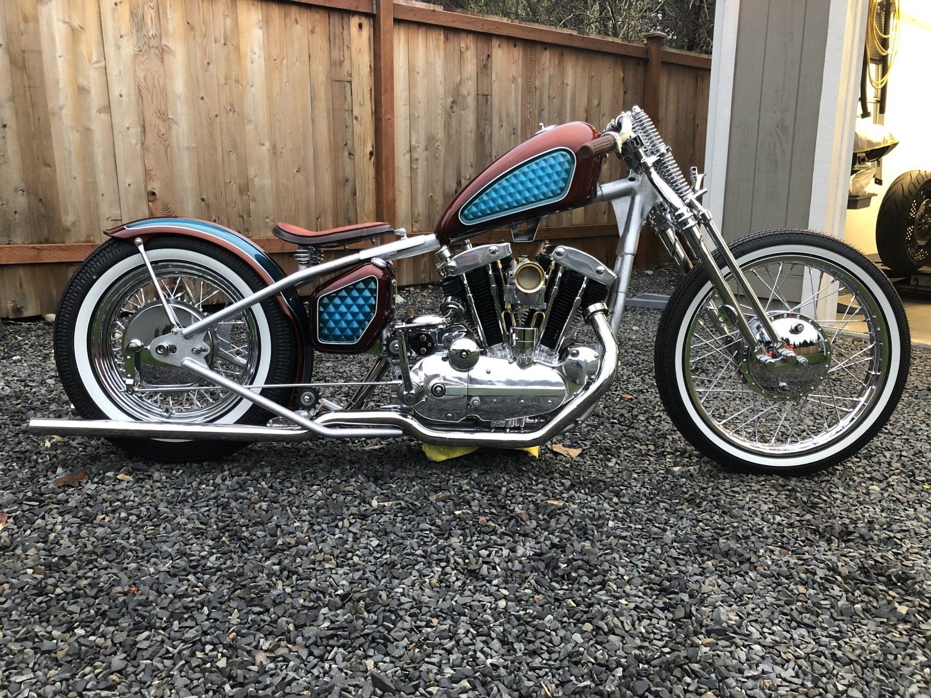 Custom chopper motorcycle with red and blue accents parked on gravel beside a wooden fence