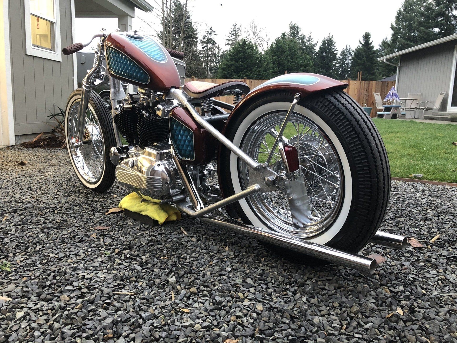 Custom trike motorcycle with red tank and chrome wheels parked on gravel beside a house.