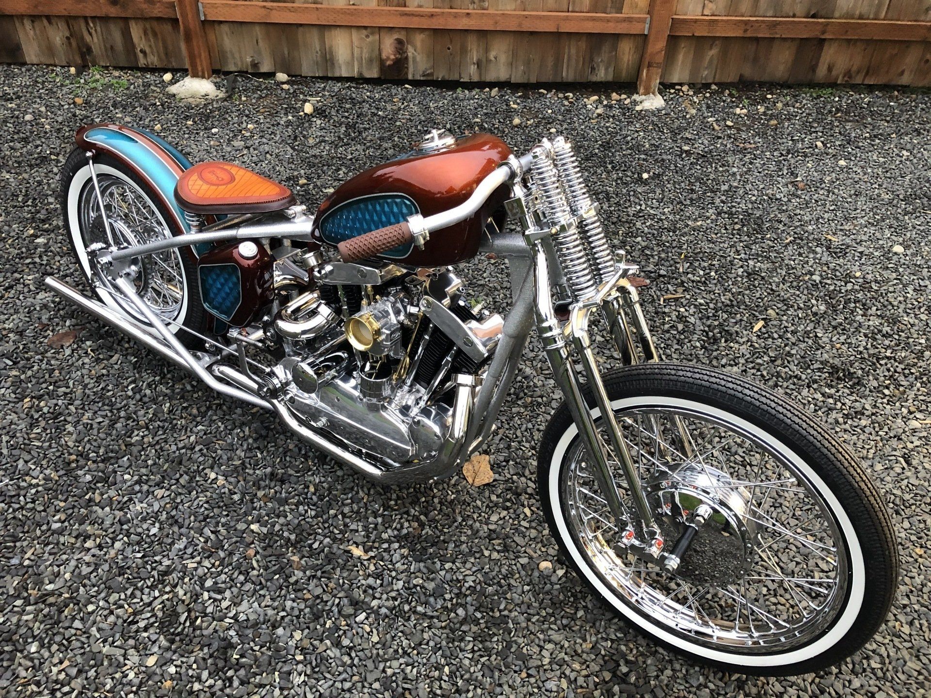 Custom chopper motorcycle with chrome details, orange seat, and whitewall front tire on gravel.