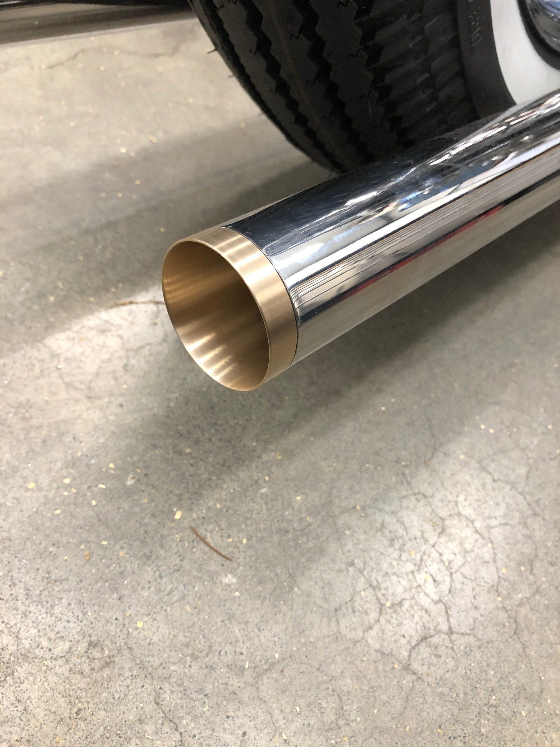 Chrome exhaust pipe tip with brass-colored inner edge beside a tire on a concrete floor