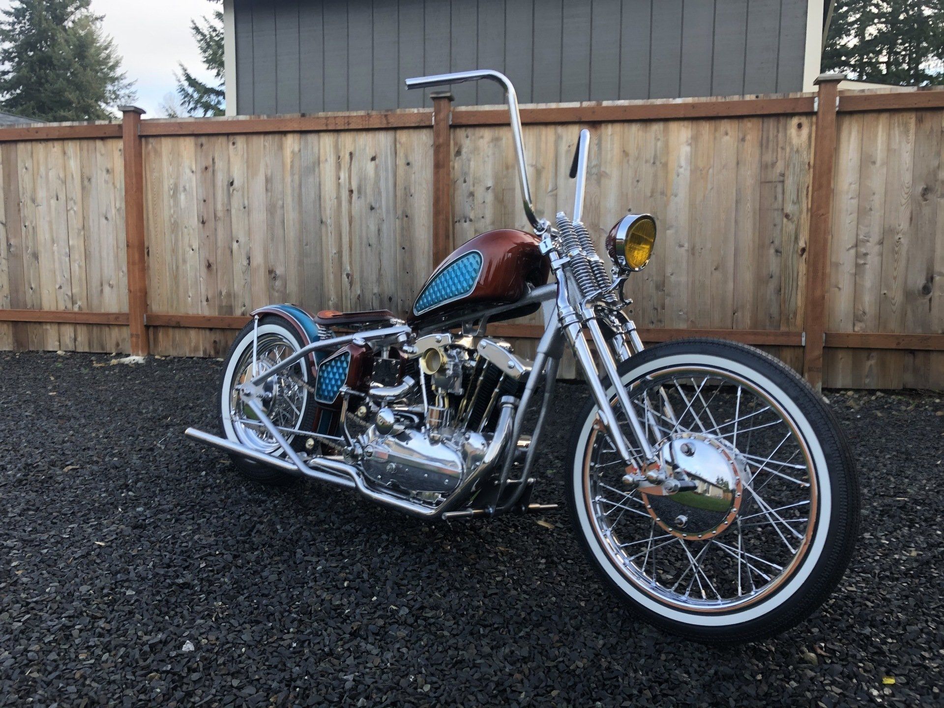 Custom chrome chopper motorcycle parked on gravel beside a wooden fence, with red tank and tall handlebars