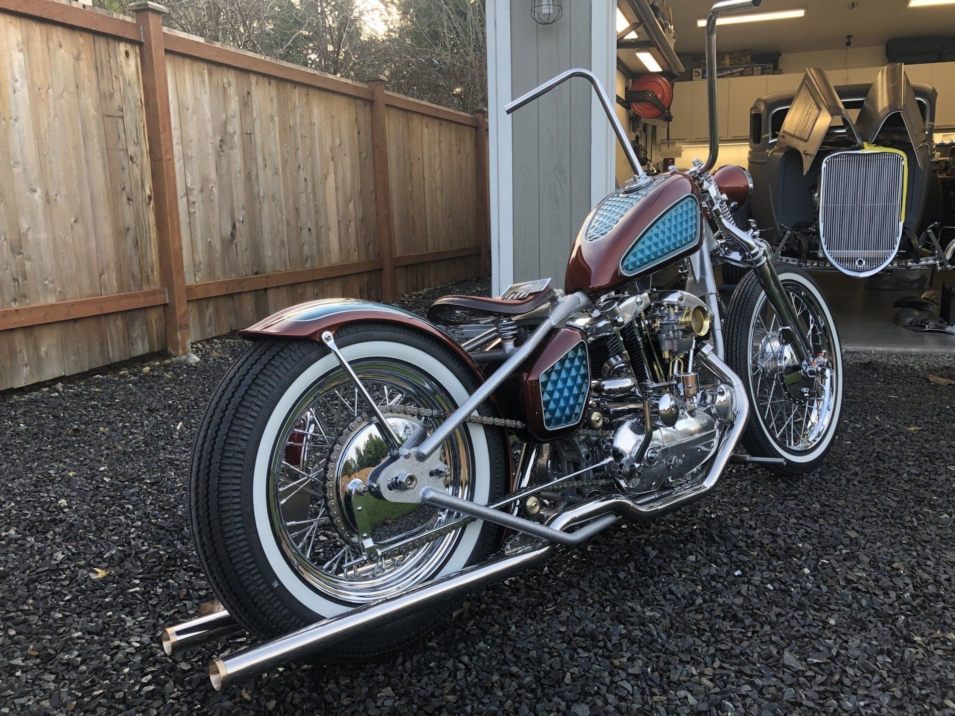 Custom chopper motorcycle parked on gravel beside a wooden fence and yellow backhoe.