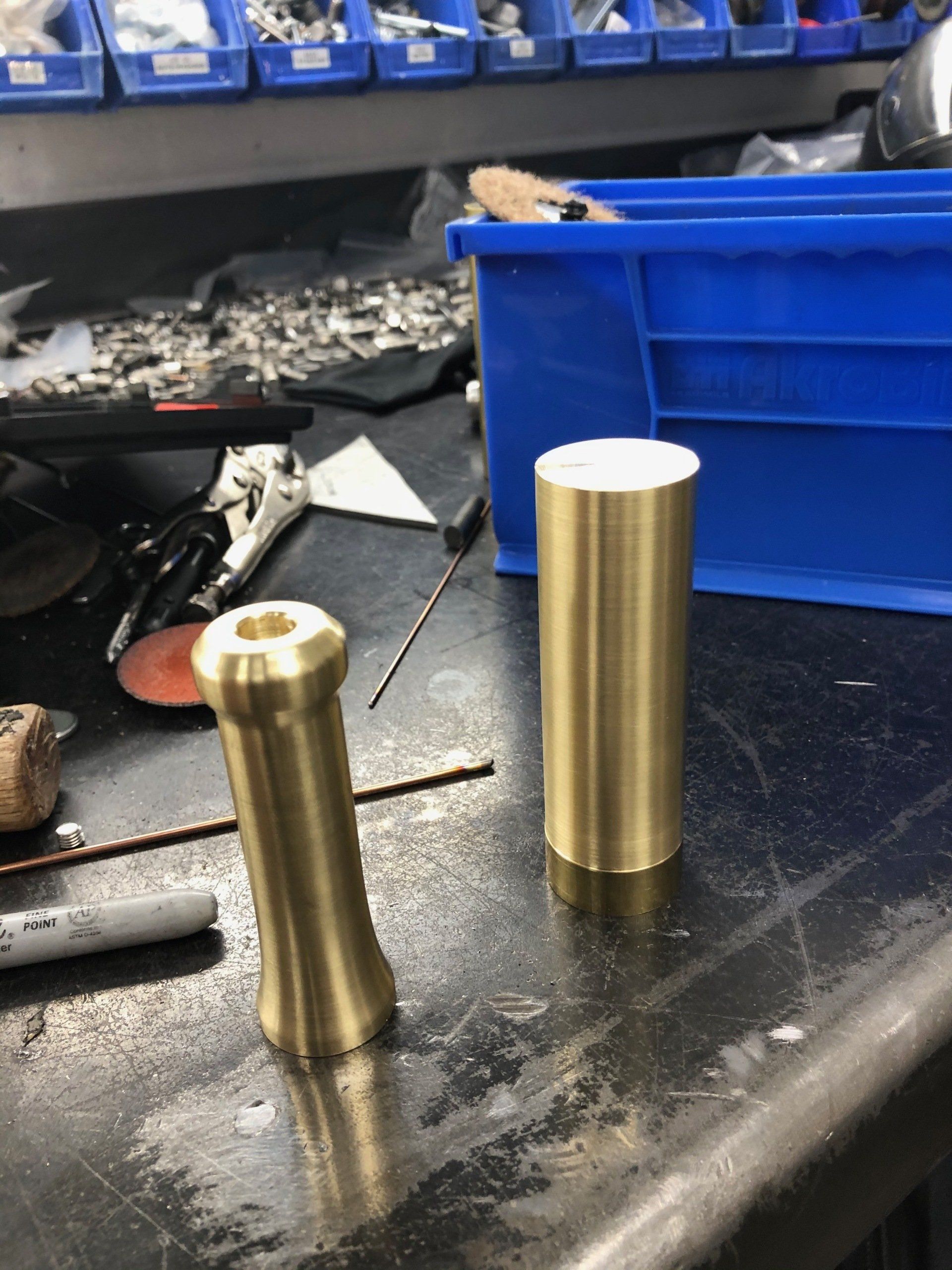 Two brass cylinders on a workshop bench, one short and worn, one tall and polished.
