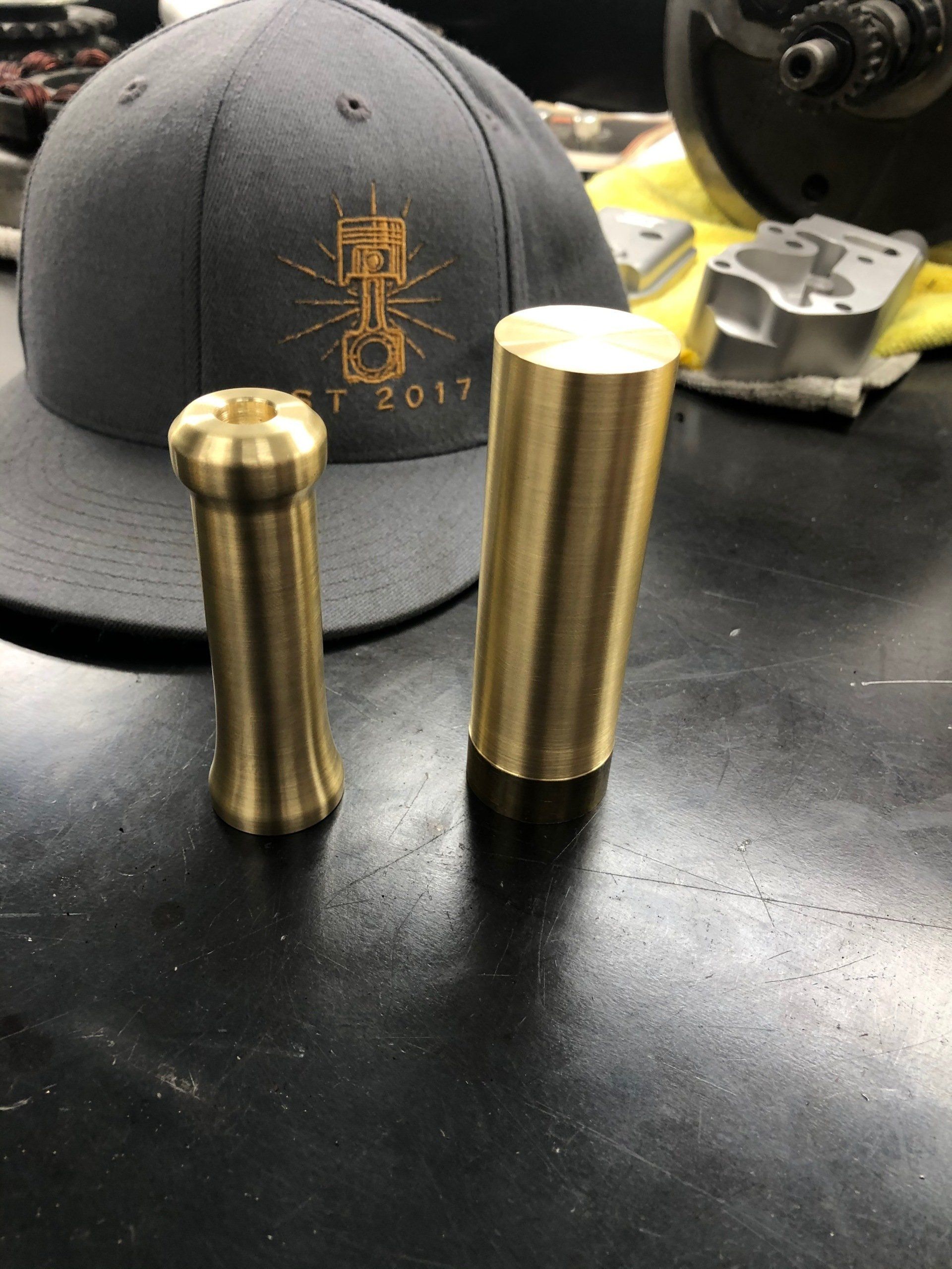 Two brass cylinders on a black workbench, with a gray cap and machinery in the background